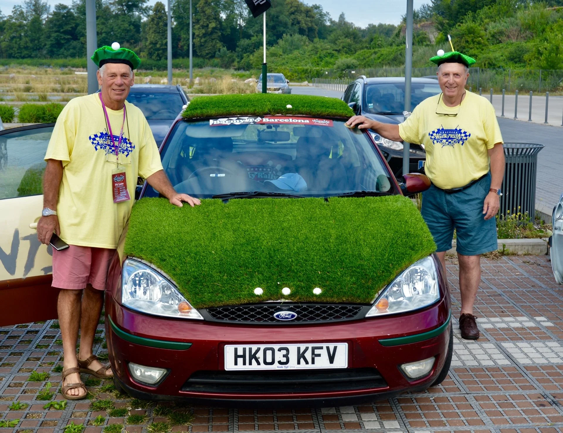 Two men wearing yellow T-shirts with 'Wacky Rally' logo, green hat decorations, and shorts, standing next to a maroon car decorated with green grass-like material on the hood and roof, parked on a brick-paved area.