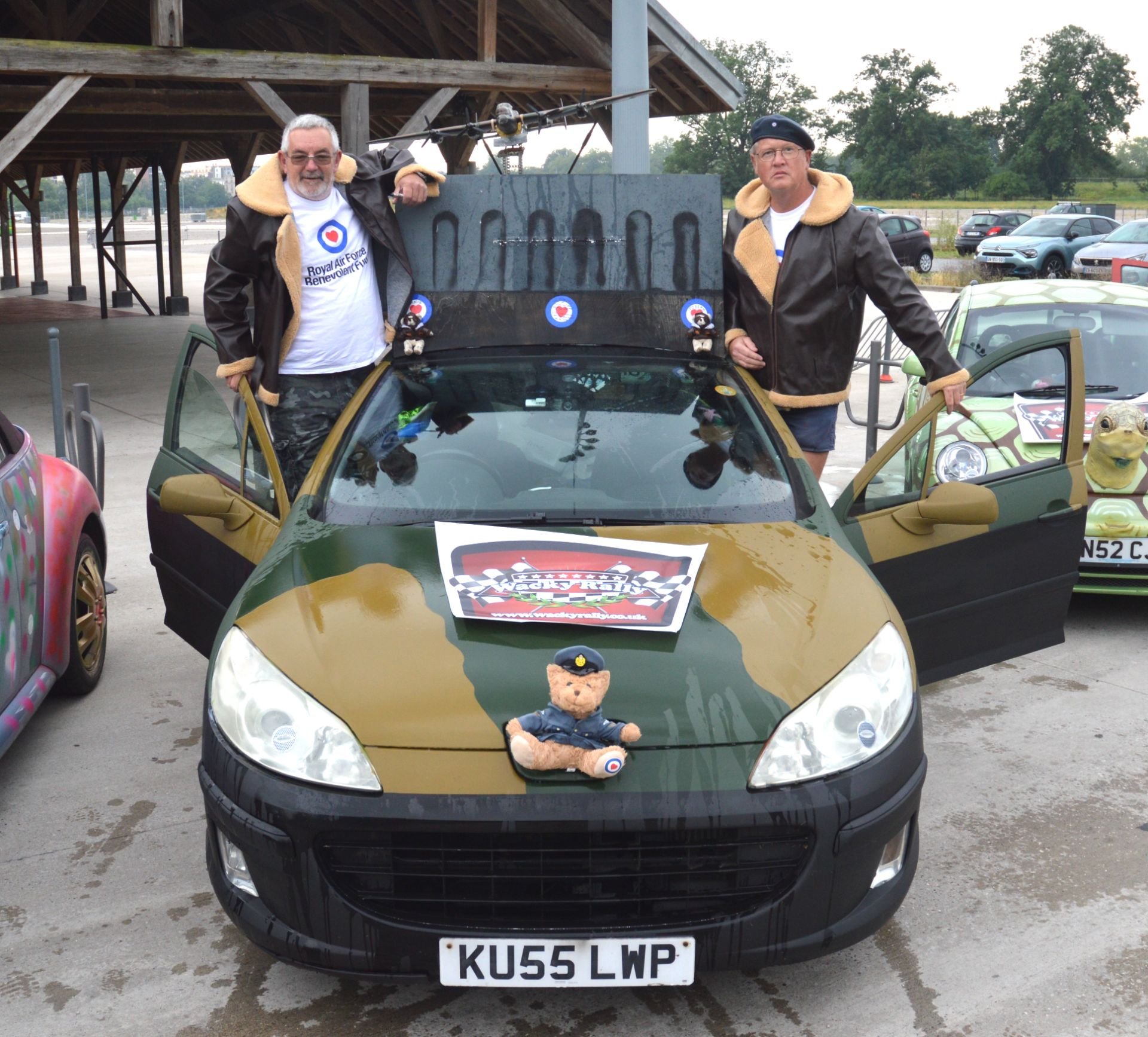 Two men stand beside a green and black sports car decorated with racing memorabilia at a car show. The car has a stuffed bear in a police uniform on the hood and a pop-up roof with military-themed insignia. One man is leaning on the open driver's sid