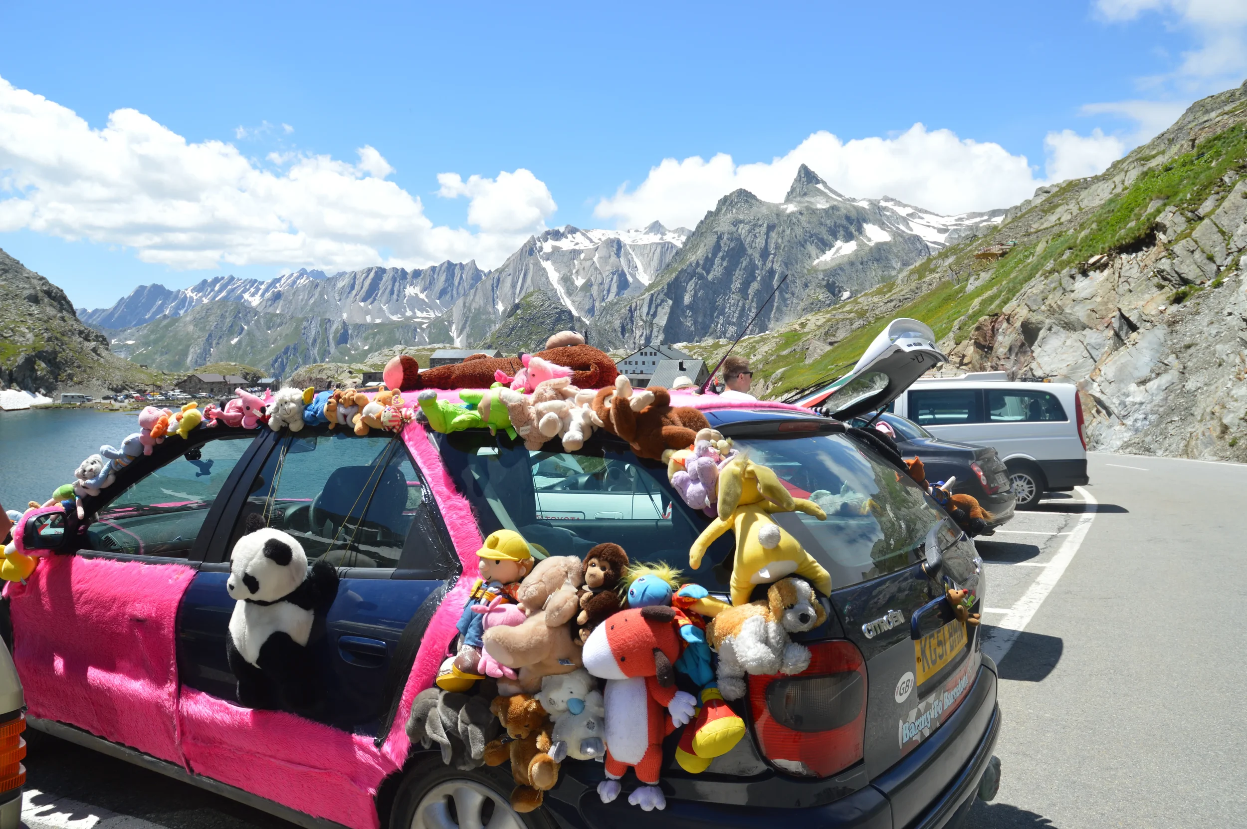 Car covered with numerous stuffed animals, parked in a mountainous area with a lake and snow-capped peaks in the background.