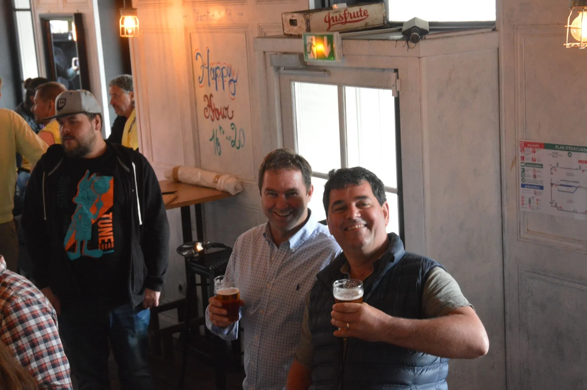 Two men smiling at a bar or pub, holding drinks, with a window behind them and a neon emergency exit sign above. Other patrons and seating areas are visible in the background.