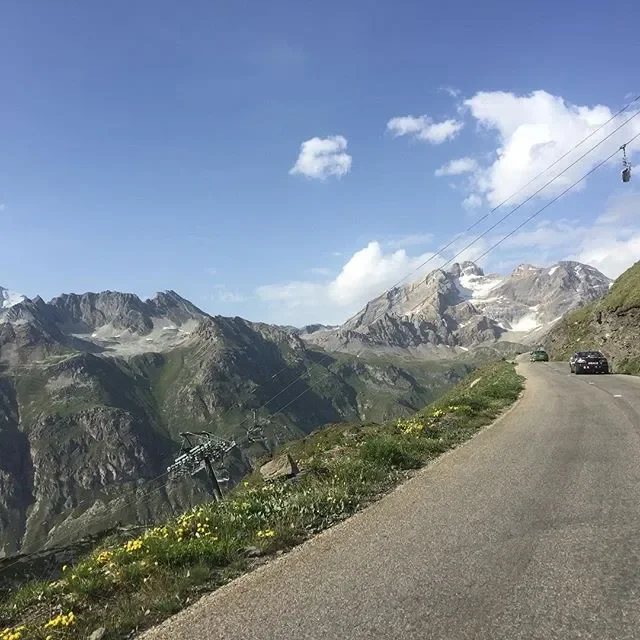 A mountain road with a car parked on the right side, surrounded by rugged mountains with patches of snow, green vegetation, and a few clouds in the sky.