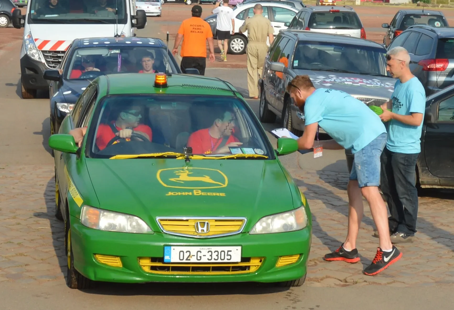 A green John Deere-themed car at a race event with three people inside and three people outside, one holding a clipboard, in a parking lot filled with other cars, some bystanders walking around.