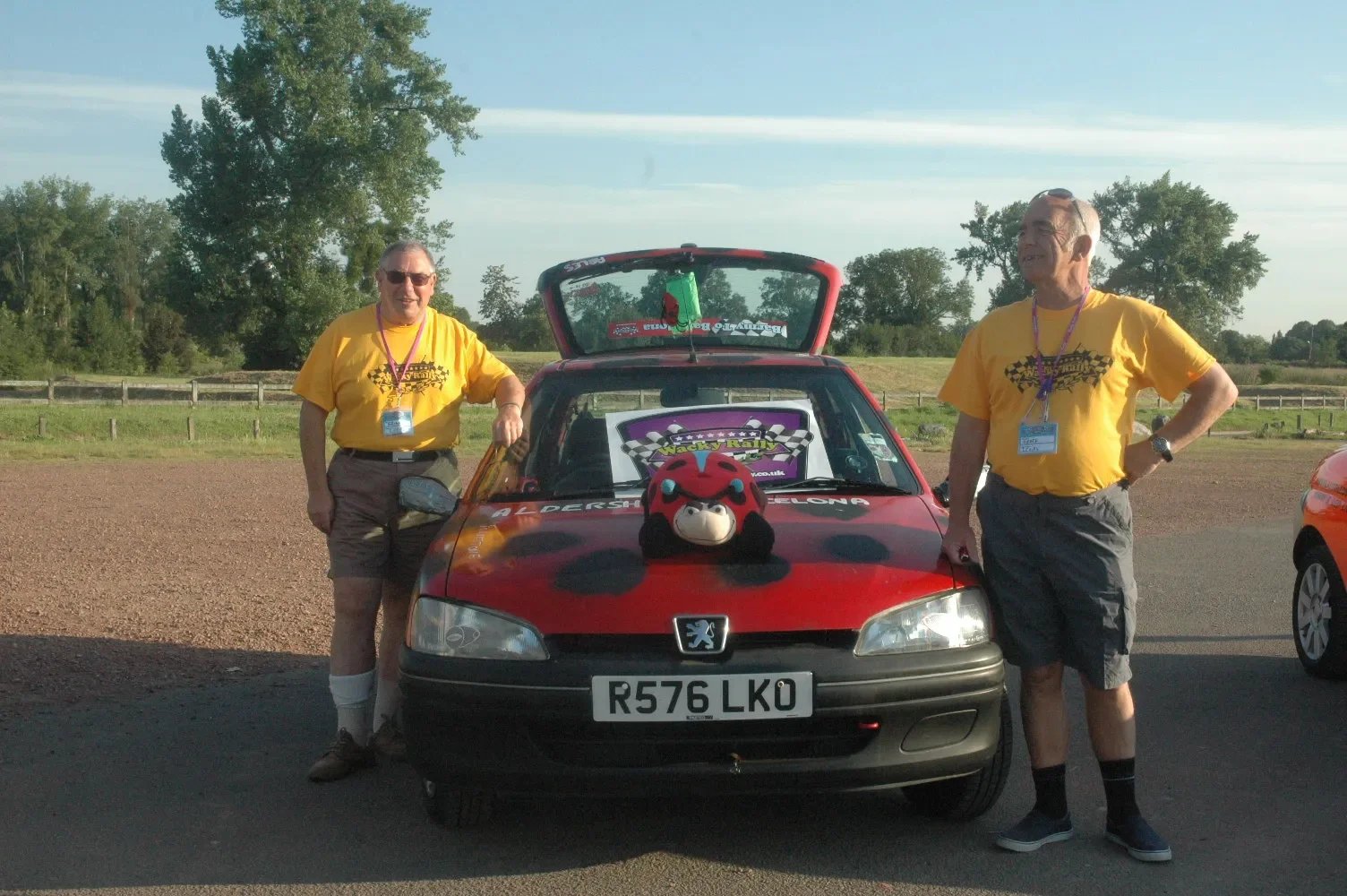 Two men standing next to a red Peugeot car at a racing event, wearing yellow t-shirts, with a plush cow toy on the car's hood, and a racing sign in the background.