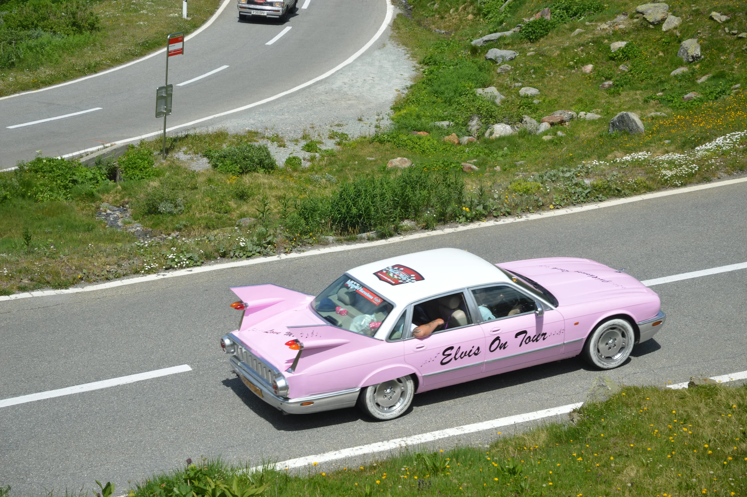 Pink vintage car with 'Elvis On Tour' written on the side traveling on a two-lane road surrounded by green grass and rocks.