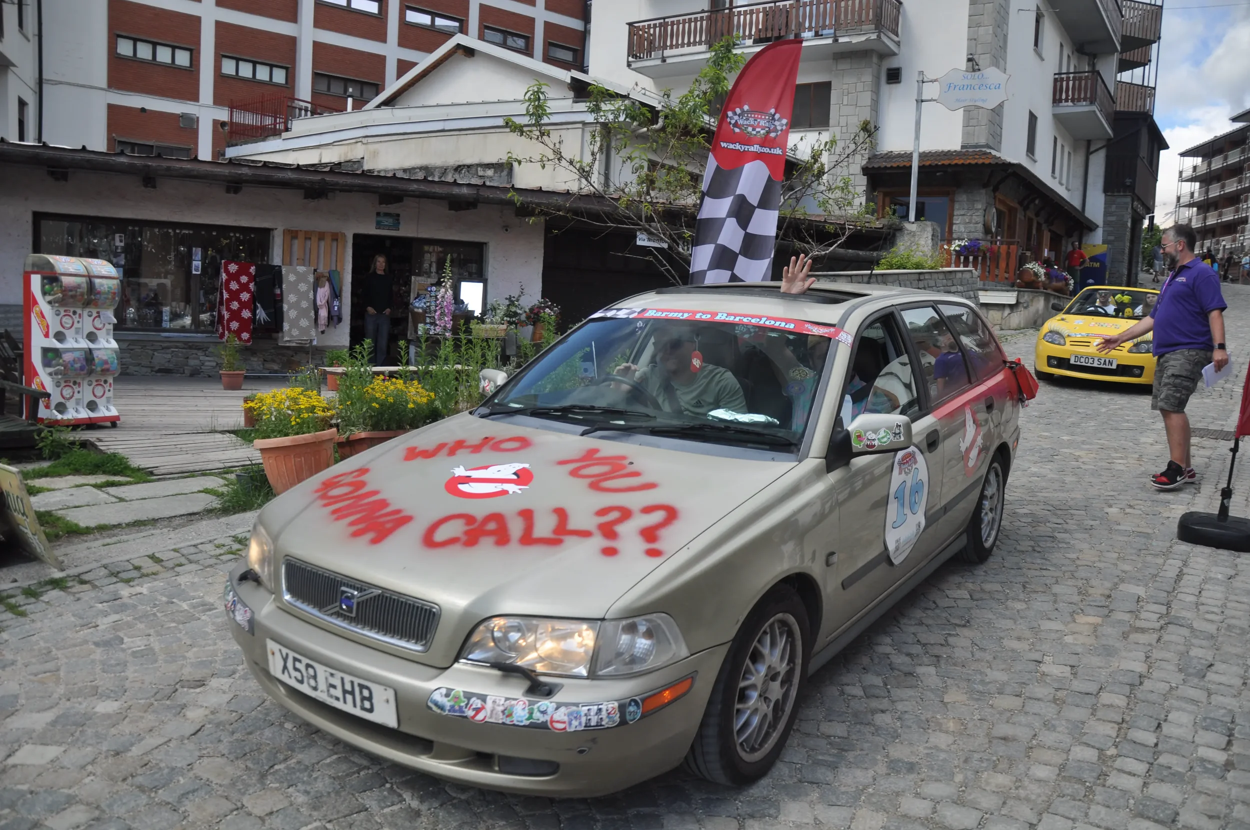 A beige Volvo car with graffiti-style writing on the hood and side, including the question "Who You Gonna Call?" and the Ghostbusters logo. The car has the number 16 on the side, a waving hand outside, and a flag on the roof. In the background, there