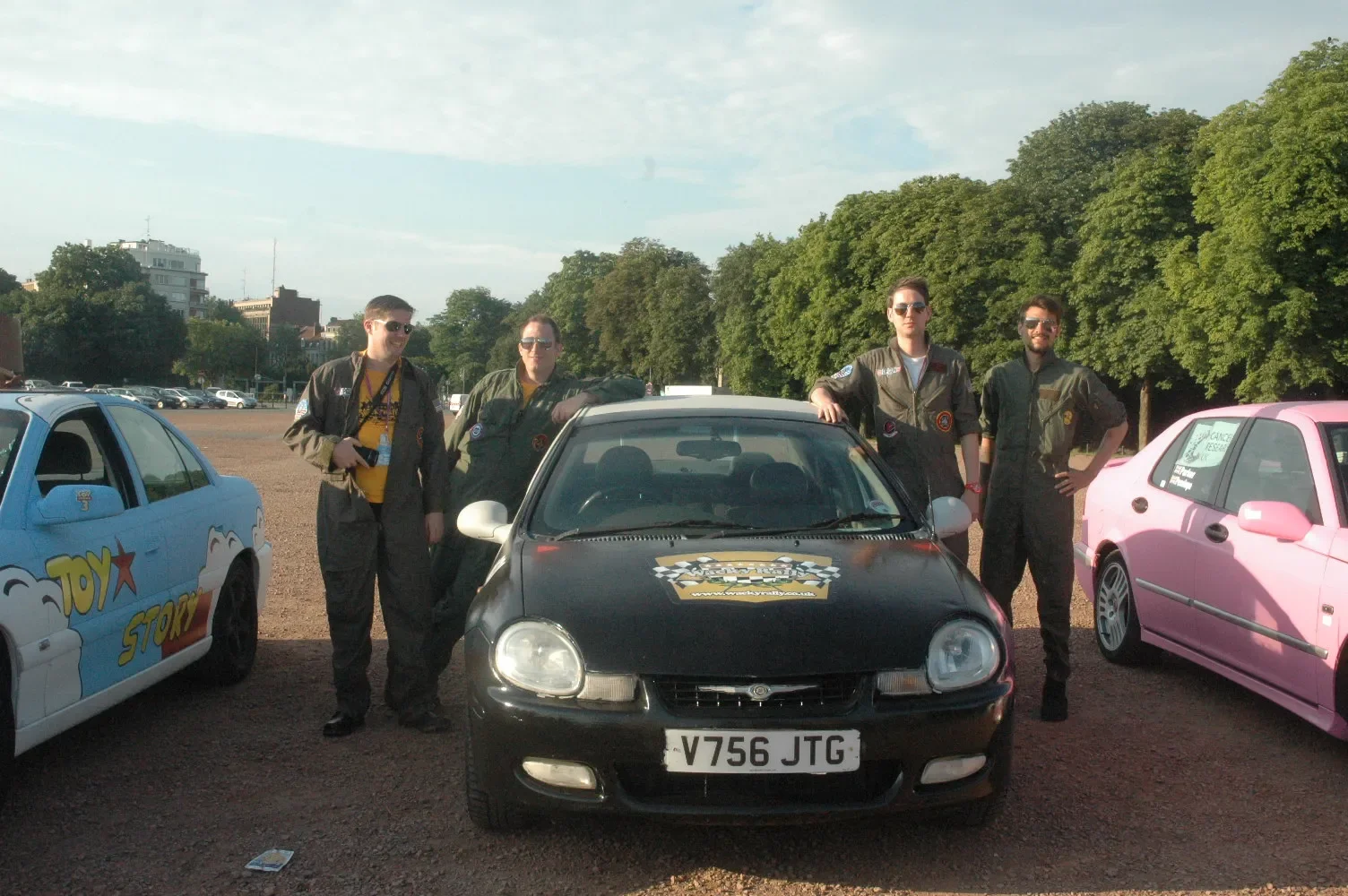 Four men in racing suits posing next to a black racing car with a checkered flag sticker on the hood, in a parking lot with modified cars, trees, and a few buildings in the background during daytime.