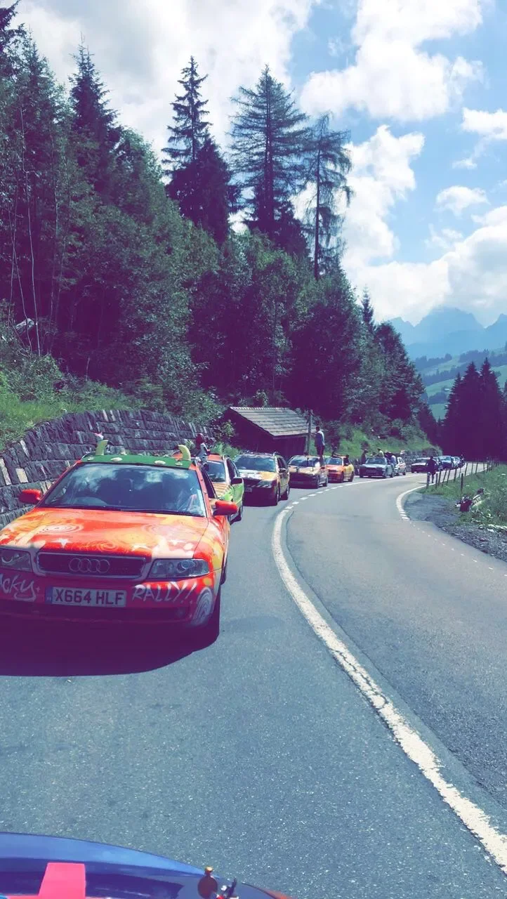 A line of cars parked along a winding mountain road with tall pine trees and a blue sky in the background.