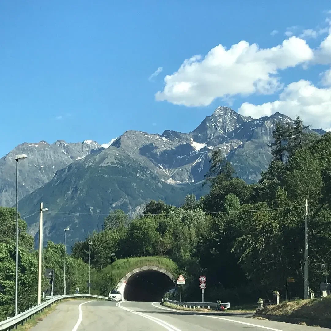 Mountain landscape with snow-capped peaks, a clear blue sky with a few clouds, and a highway tunnel entrance surrounded by green trees.