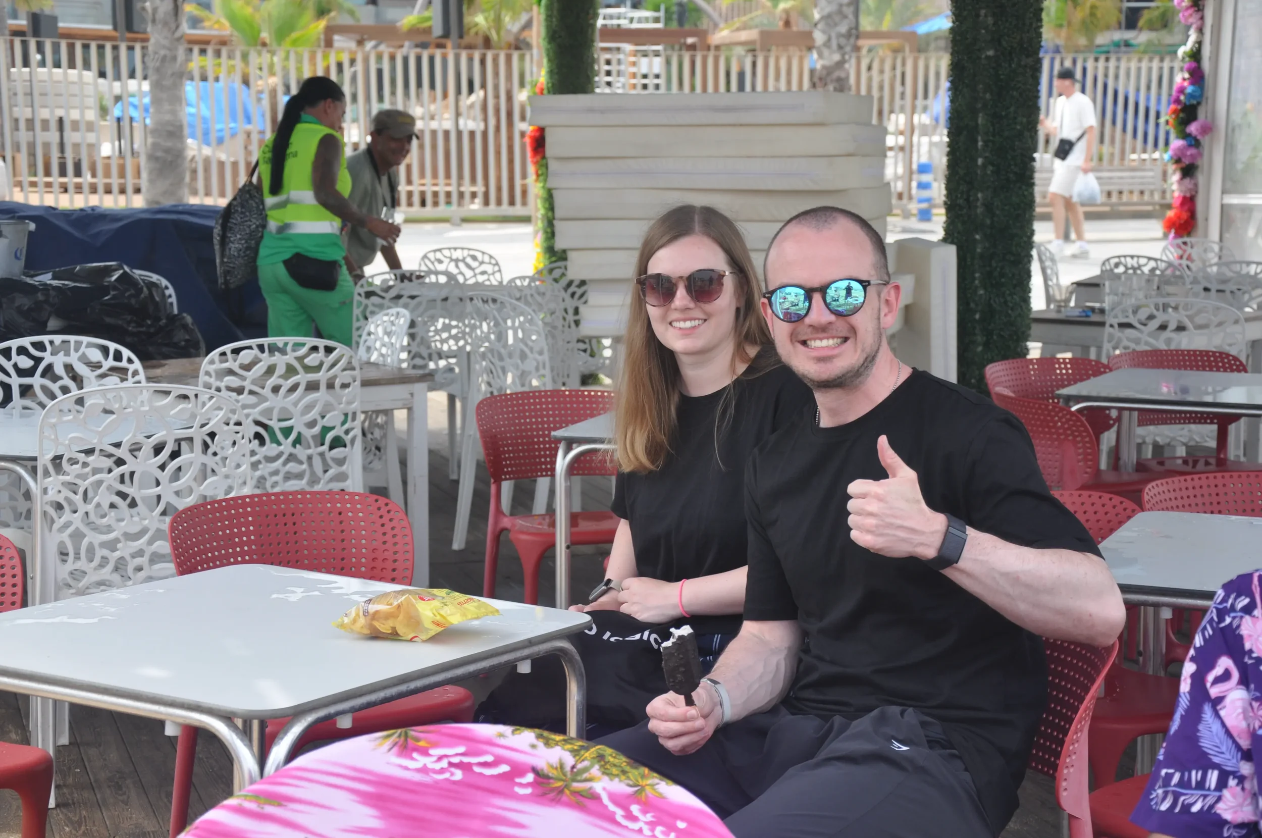A smiling young man and woman wearing sunglasses sitting outdoors at a cafe, with the man giving a thumbs-up and holding a partially eaten ice cream bar. There are empty chairs and tables around them, and a woman in a green safety vest and another pe