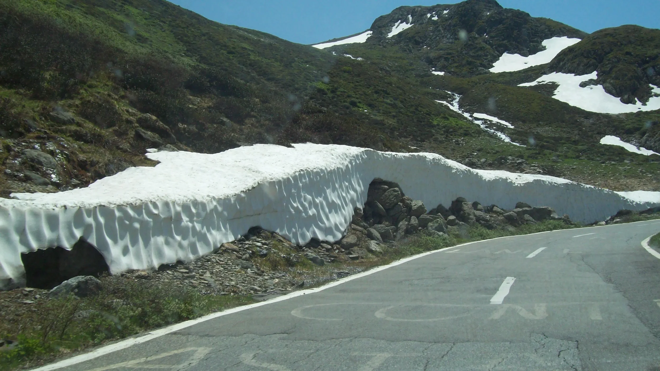A mountain road with a sharp curve, with a snowbank on the side that extends over the edge and onto the road. The snowbank is large and uneven, with rocks at its base. The mountain in the background has patches of snow and green vegetation.