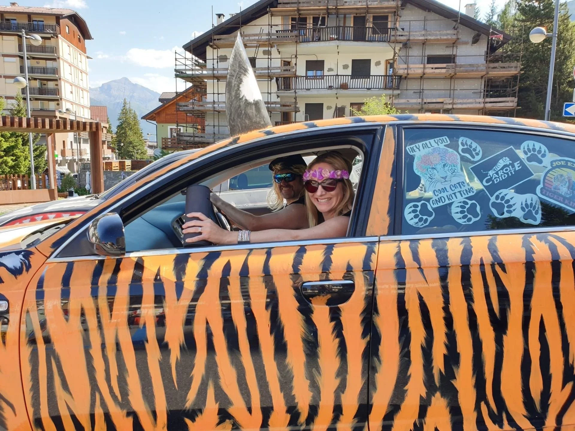 A woman and a man inside a car with a tiger stripe pattern, the woman is wearing sunglasses and a crown of purple flowers, and the man is wearing sunglasses, with a backdrop of buildings and mountains.