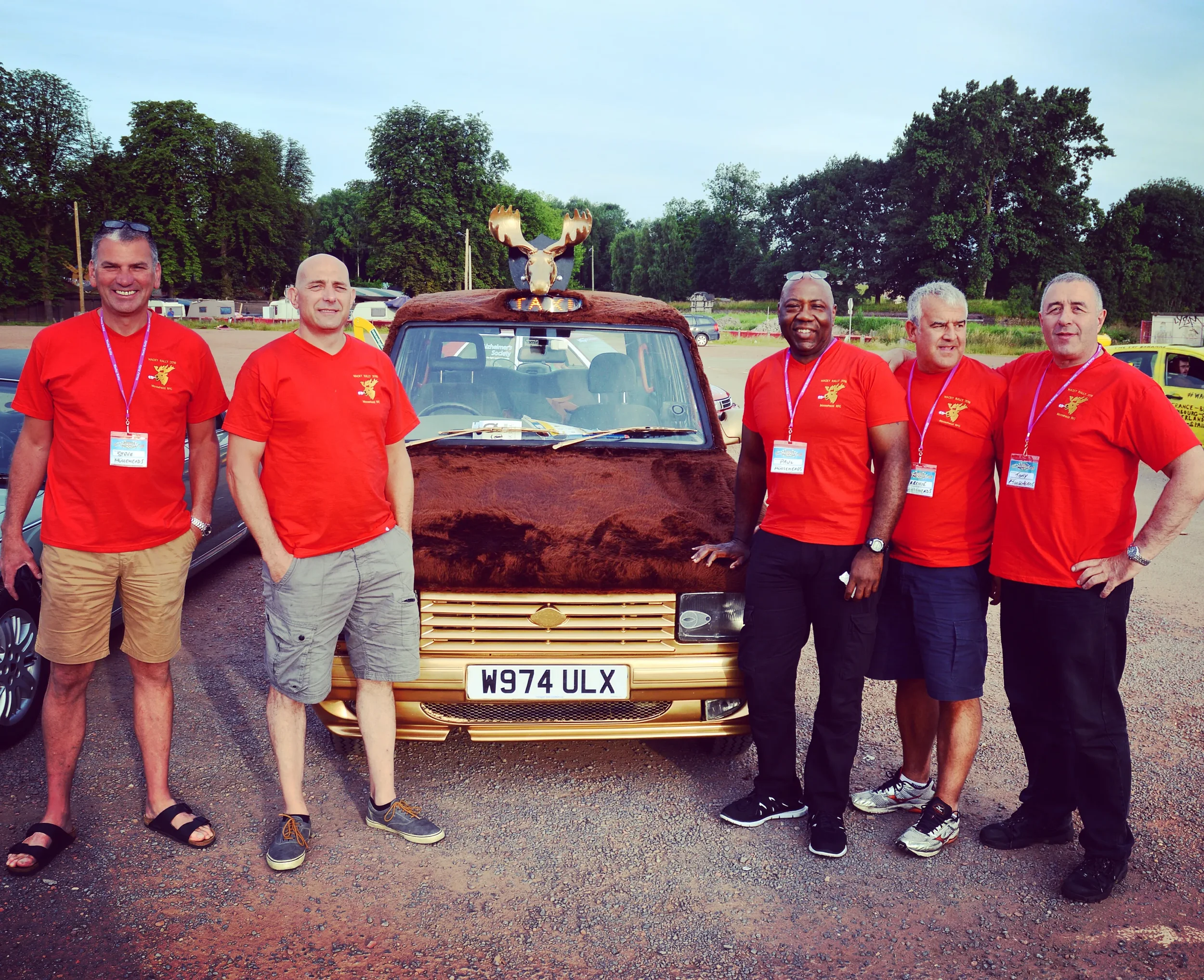 Six men standing in front of a decorated gold car with a moose head on the roof. They are at an outdoor event, all wearing red shirts with a logo and name tags.
