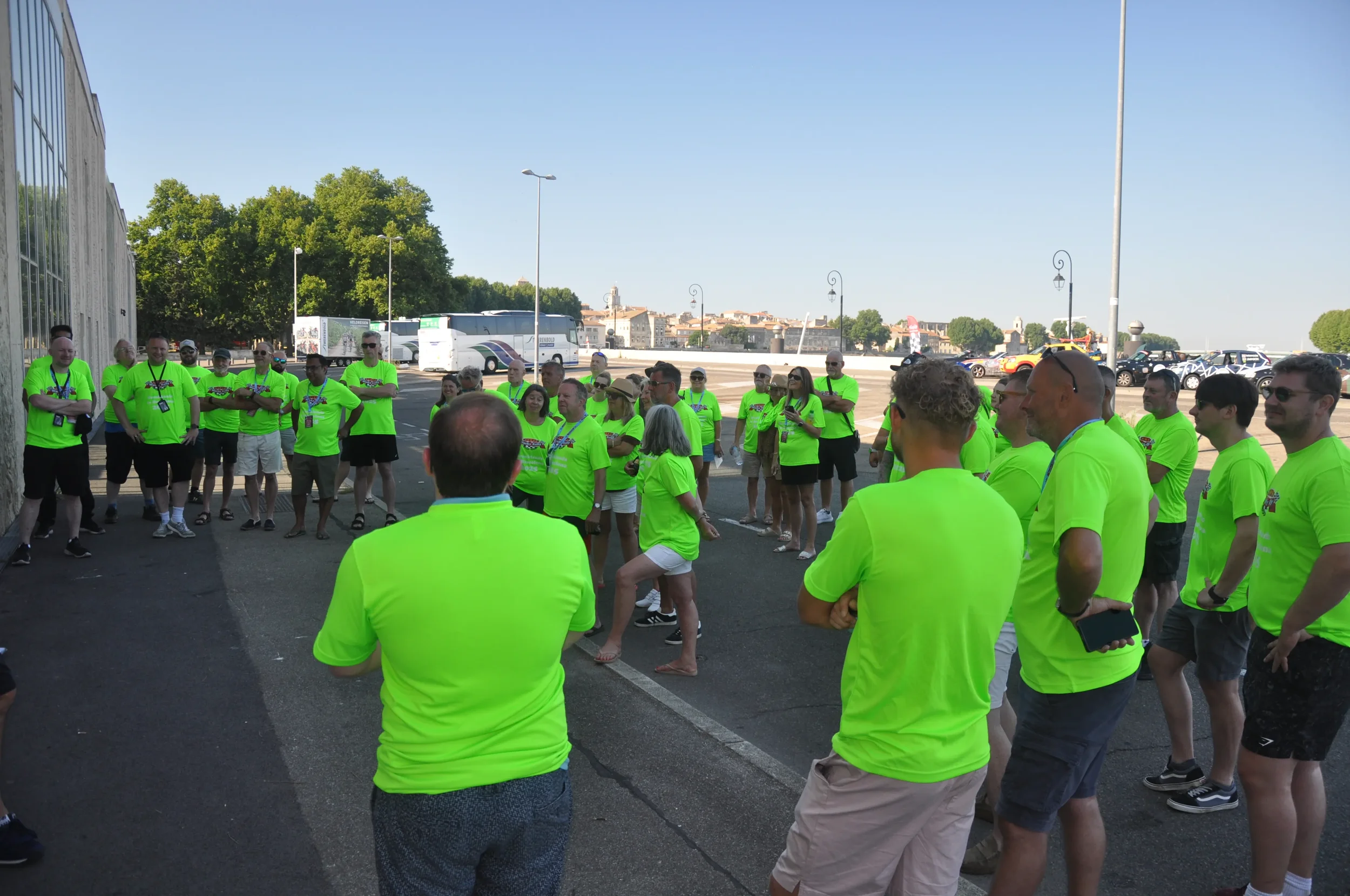 Group of people wearing matching bright green T-shirts gathered outdoors for a meeting or event. Some are listening while a person in front appears to be speaking.