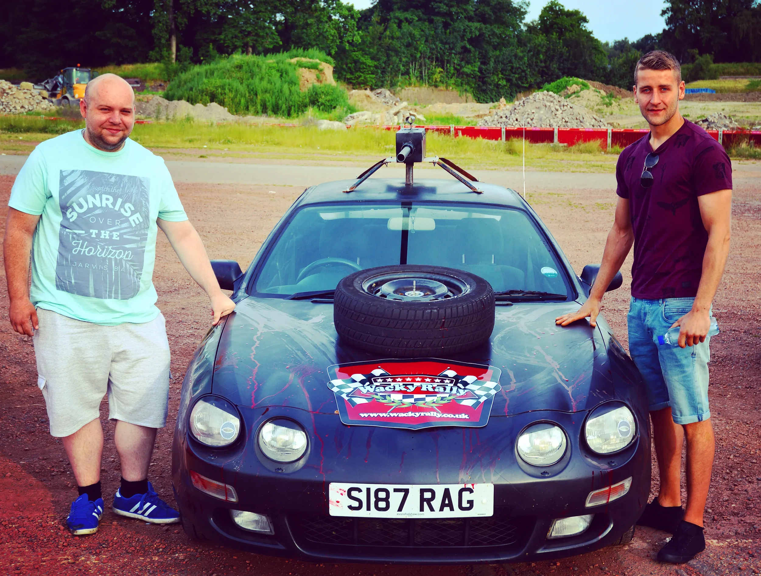 Two men standing next to a black sports car with a spare tire on the hood, in an open outdoor area with construction materials and green trees in the background.