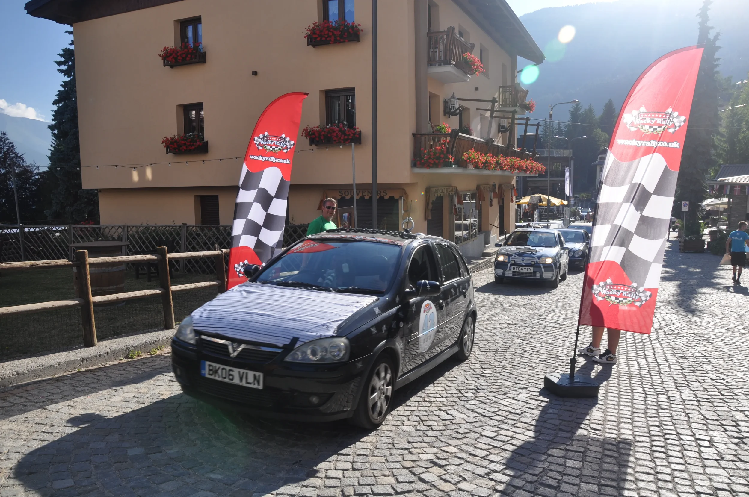 A black car with race stickers and a concealed front hood participating in a street race event, flanked by red checkered flags and people observing on a cobblestone street in a mountainous town with buildings and outdoor cafes.