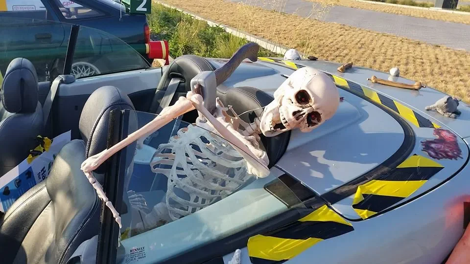 A skeleton skeleton is sitting in the driver's seat of a car with its head leaning back and mouth open. The skeleton's arm is resting on the steering wheel. The car's hood is decorated with black and yellow caution tape and there are rubber rats on t