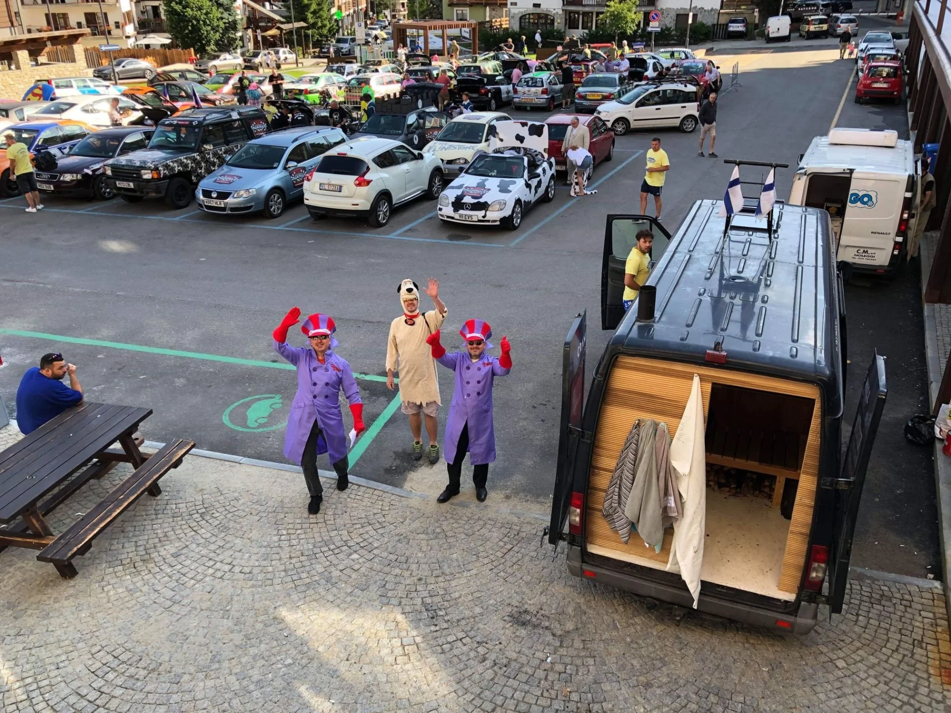 People dressed as clowns and a person in a dog costume greeting in a parking lot, with numerous cars and a food truck nearby.
