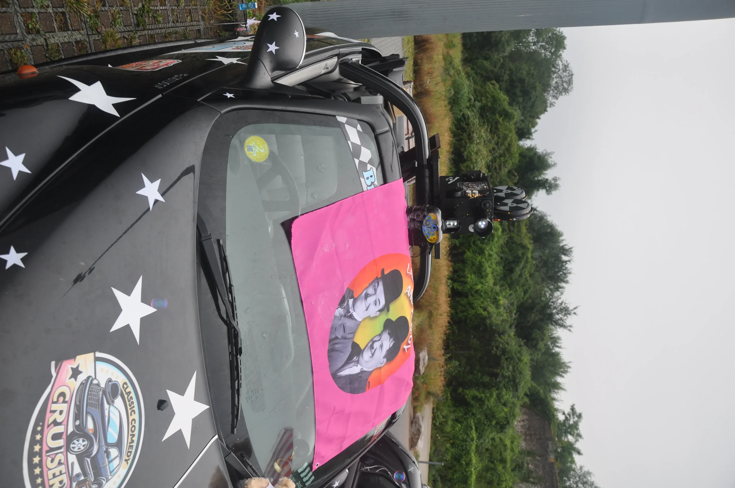 A car with star decals on the hood, a badge that reads 'GRUSE,' and a pink banner with an illustration of two women with retro hairstyles on the dashboard.
