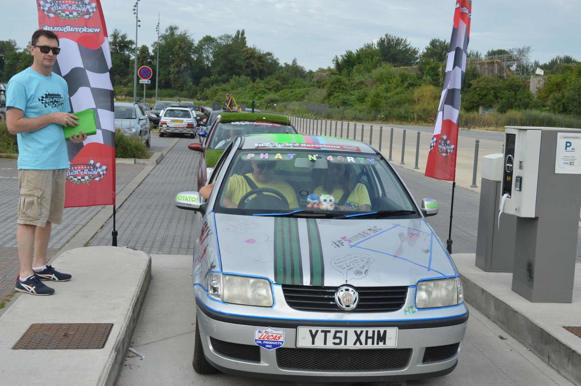 A silver MG hatchback decorated with colorful writings and drawings, parked at a race track start line with flags and a man standing next to it. Inside the car, two people are seated, wearing yellow shirts, with stuffed toys on the dashboard.