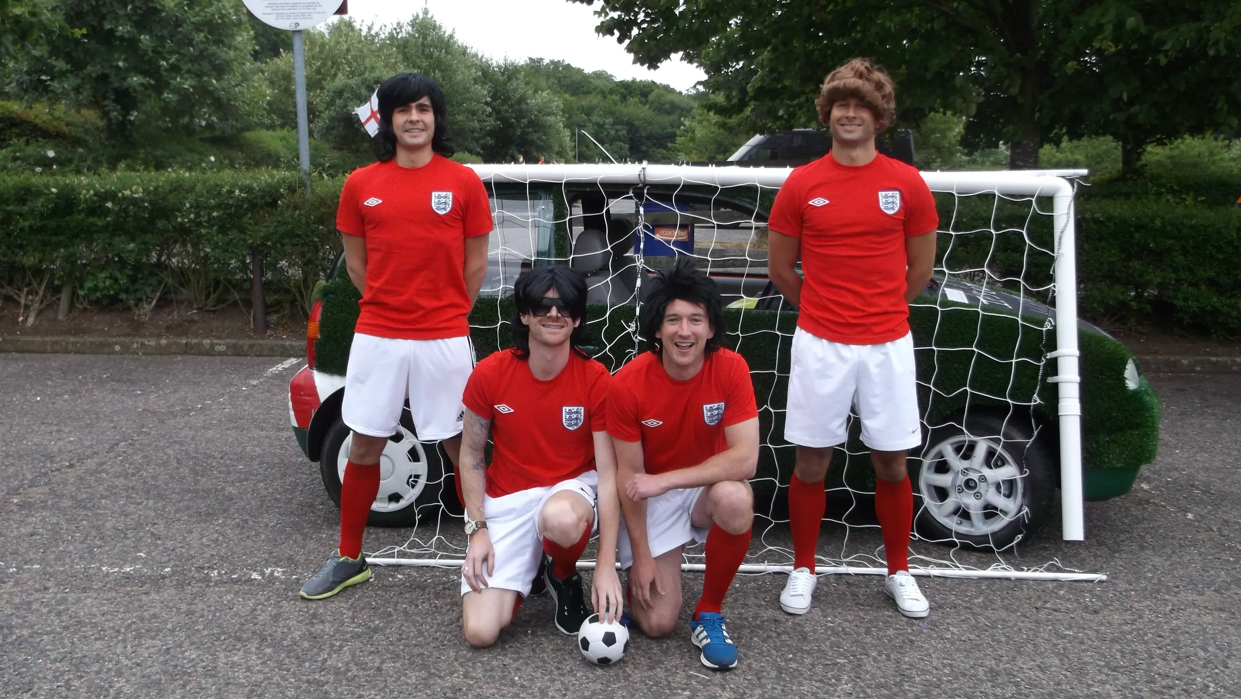 Four young men in soccer jerseys and shorts standing and kneeling in front of a mini soccer goal and a car, outdoors in a parking lot with trees in the background.