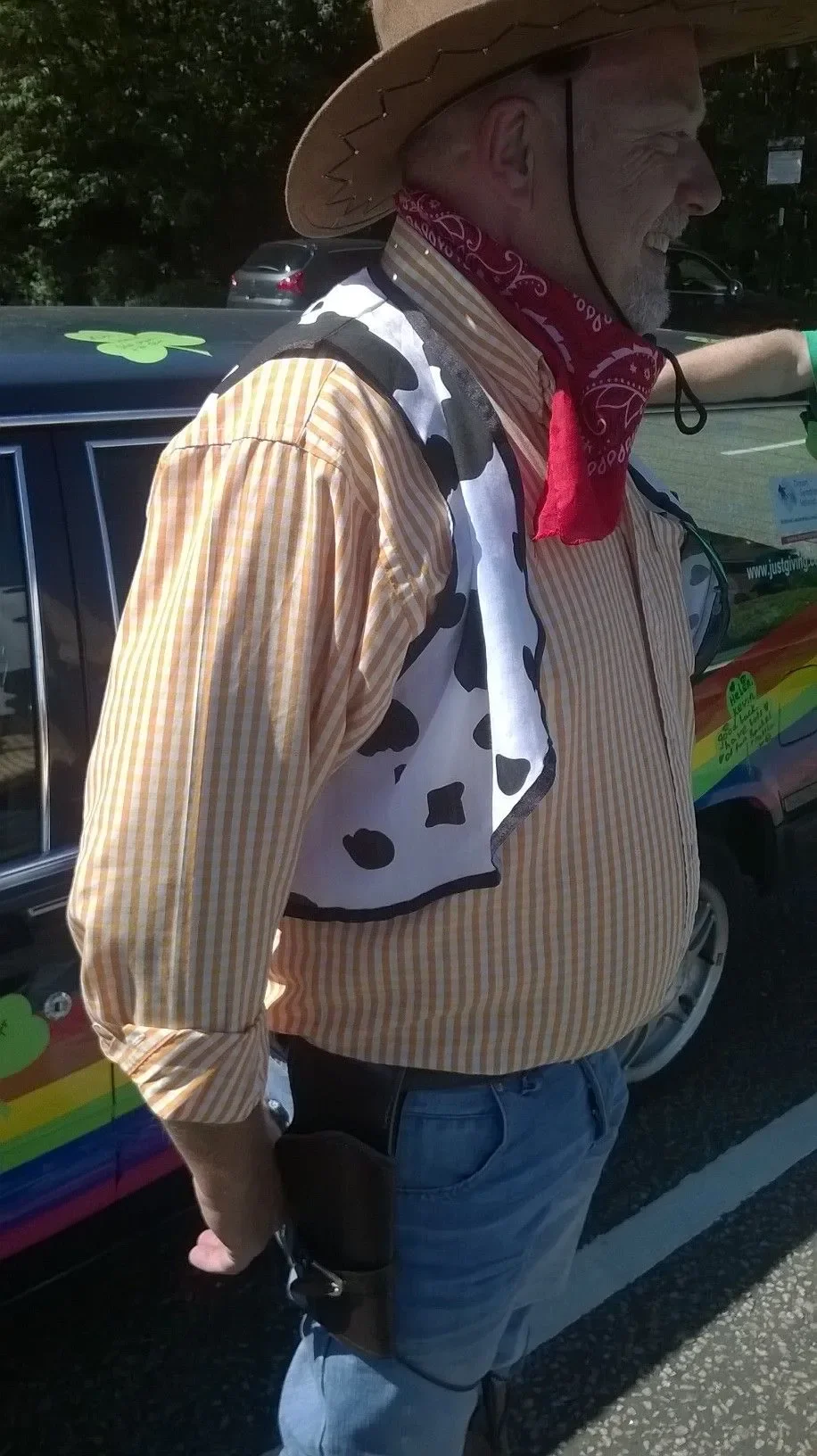 A man dressed as a cowboy with a straw hat, bandana, and cow print vest, standing next to a decorated vehicle.