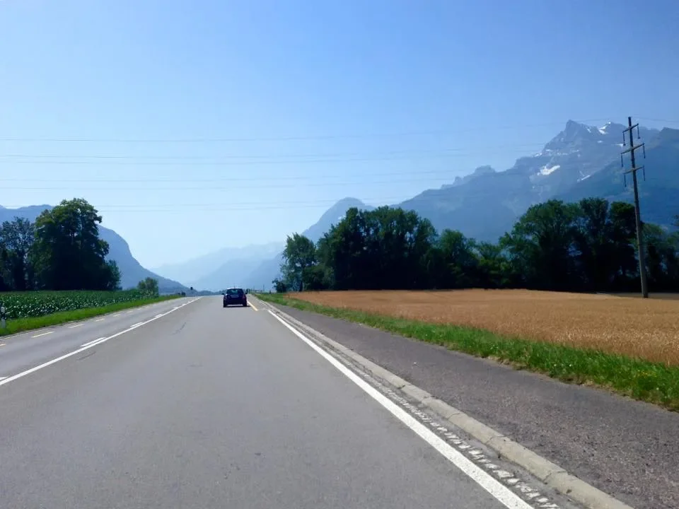 A two-lane road running through a rural landscape with mountains in the background, trees on both sides, and a car driving ahead.