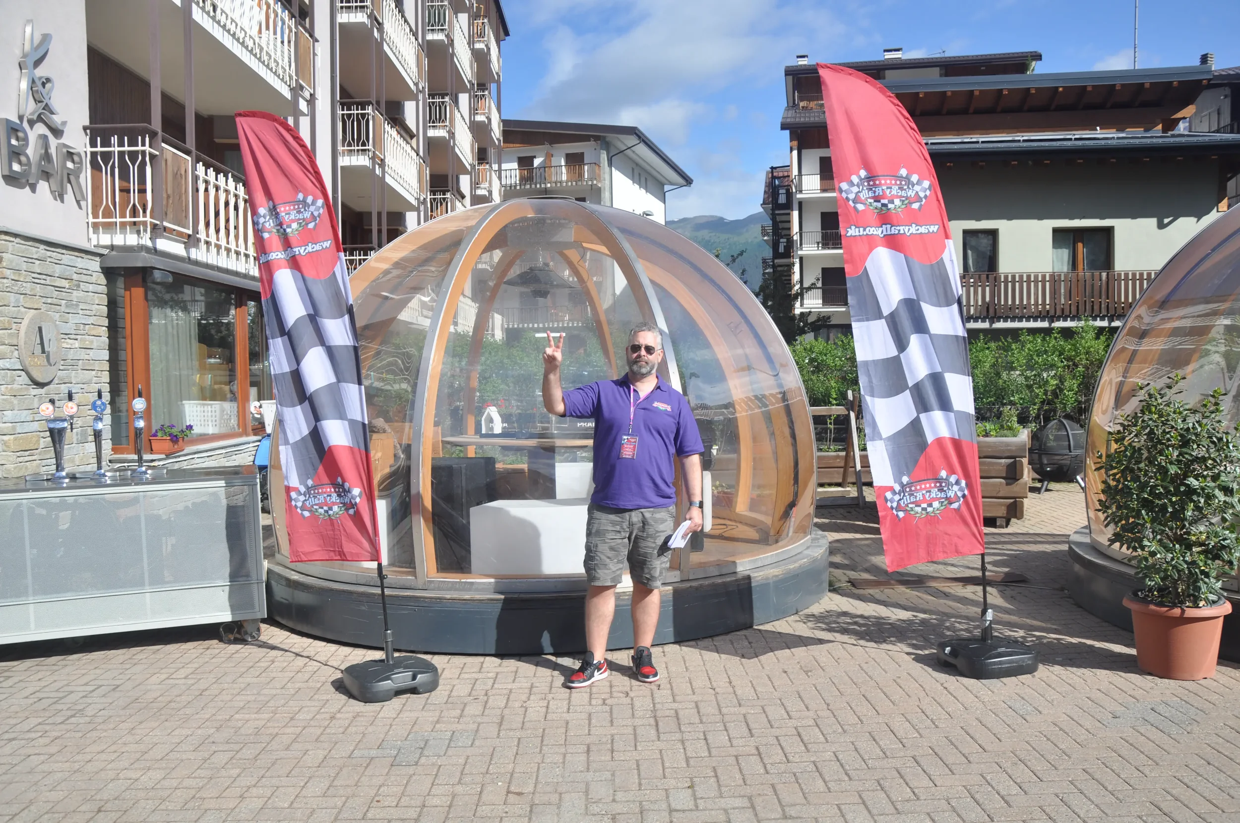 A man standing outdoors at a racing event, posing with a hand gesture, in front of a transparent dome structure with two vertical flags that have checkered flag designs and text.