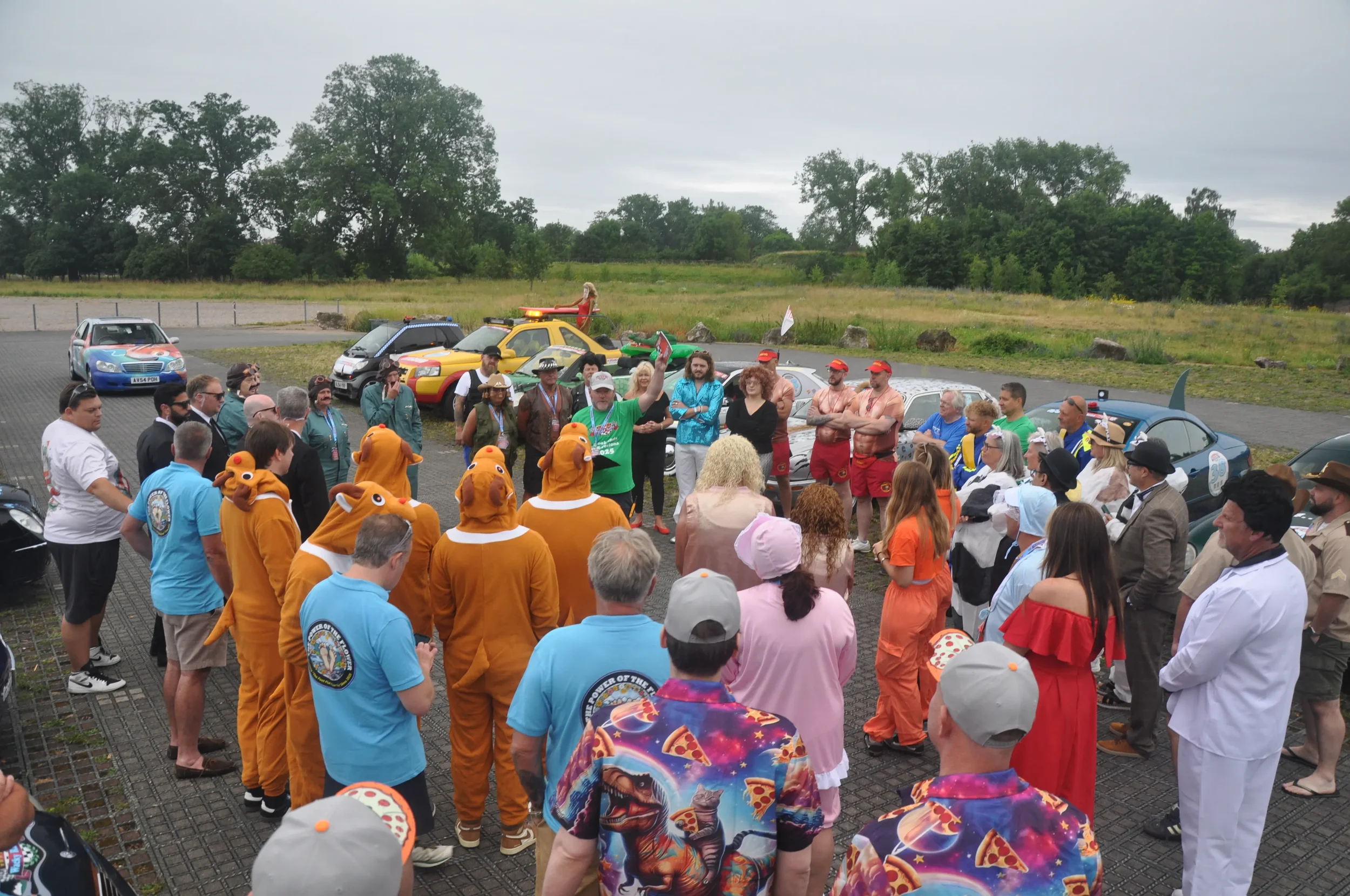 People dressed in various costumes gathered outdoors in a parking lot, with some cars and trees in the background, participating in a themed event or gathering.