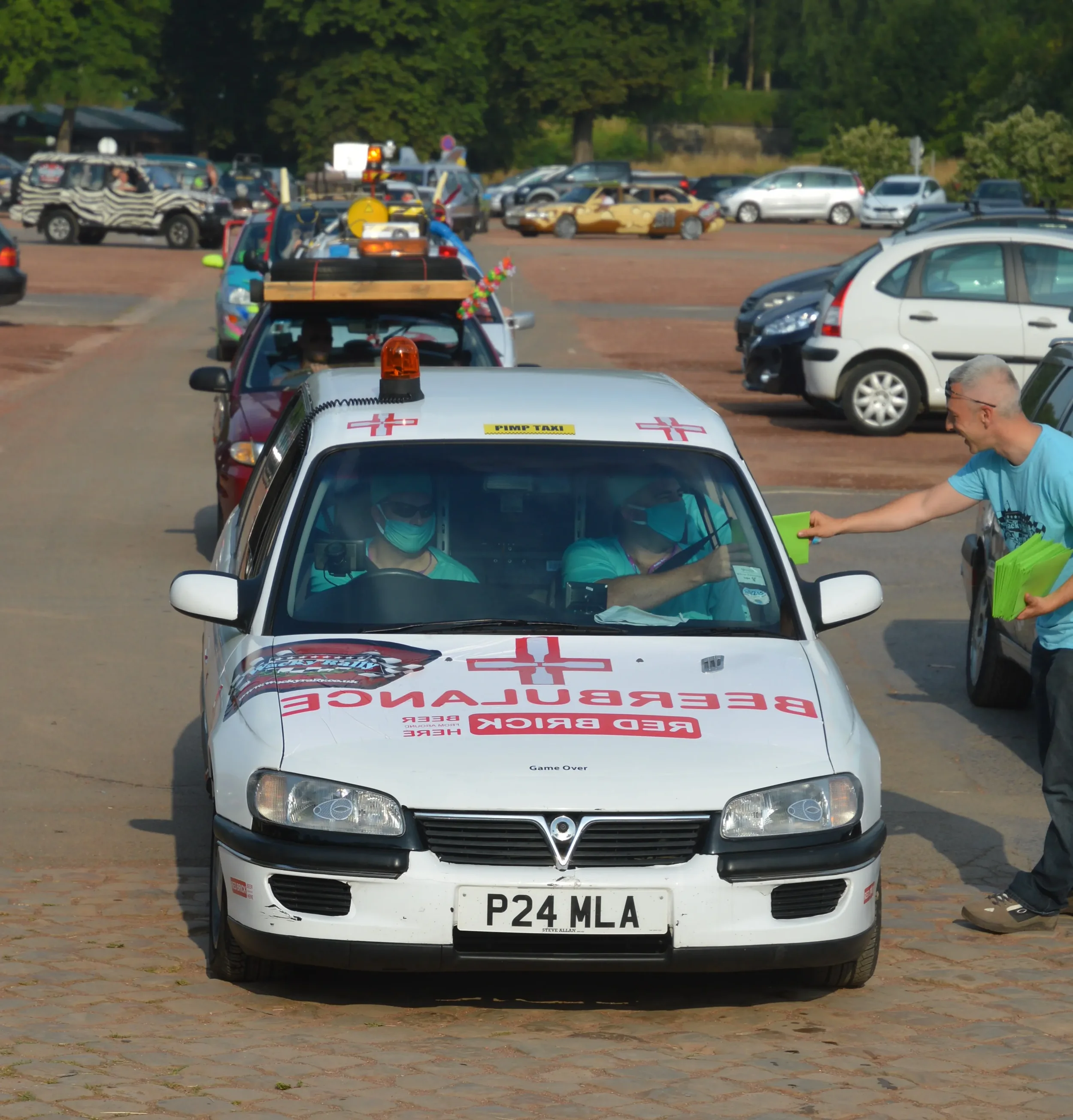 A white vehicle with medical symbols and red cross markings, serving as an emergency vehicle, parked in a lot with people interacting. Two people inside the vehicle wear masks, and a man outside is handing something green to the driver.