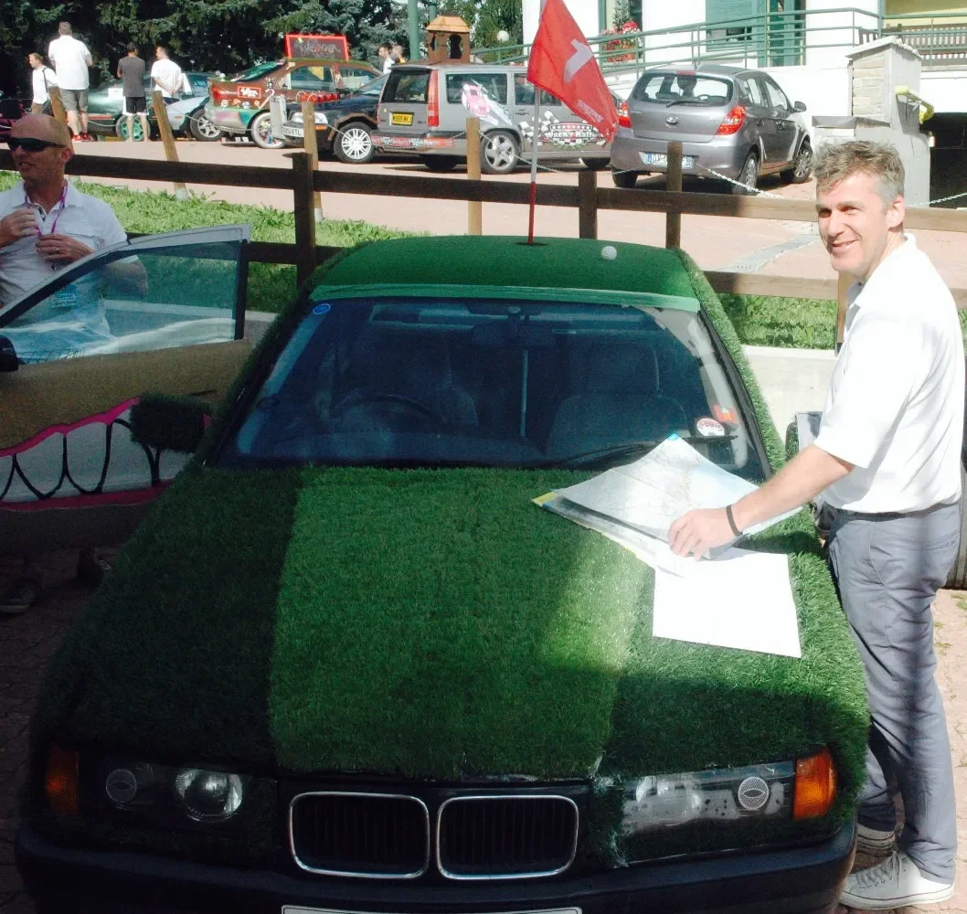 A man standing next to a BMW car covered in artificial grass, holding papers and smiling. Other people and parked cars are in the background during daytime.