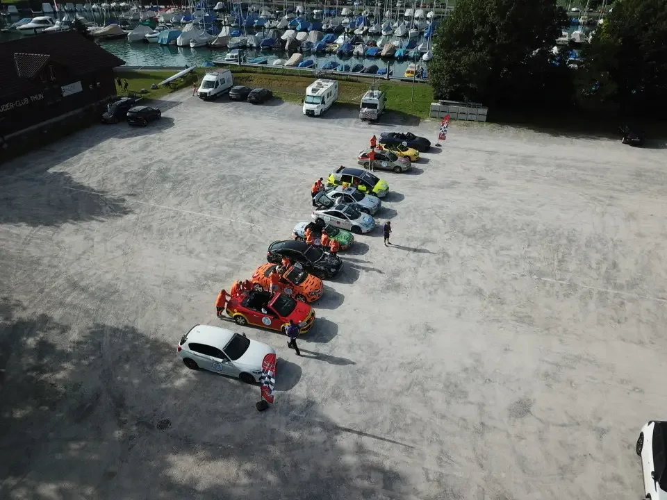 A lineup of small cars, some with racing liveries, parked in a large, open gravel lot near a marina with sailboats docked. Several people are standing near the cars, and the scene is captured from an aerial view during daytime.