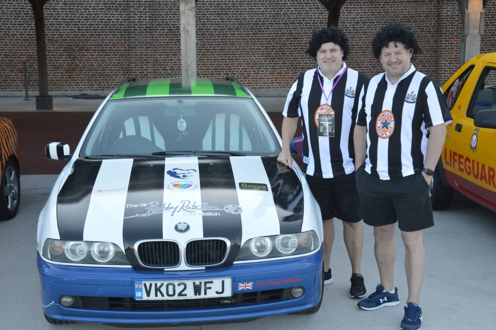 Two men dressed in black and white striped shirts with lanyards standing next to a decorated BMW car with various decals and a custom license plate, with a yellow lifeguard vehicle in the background.