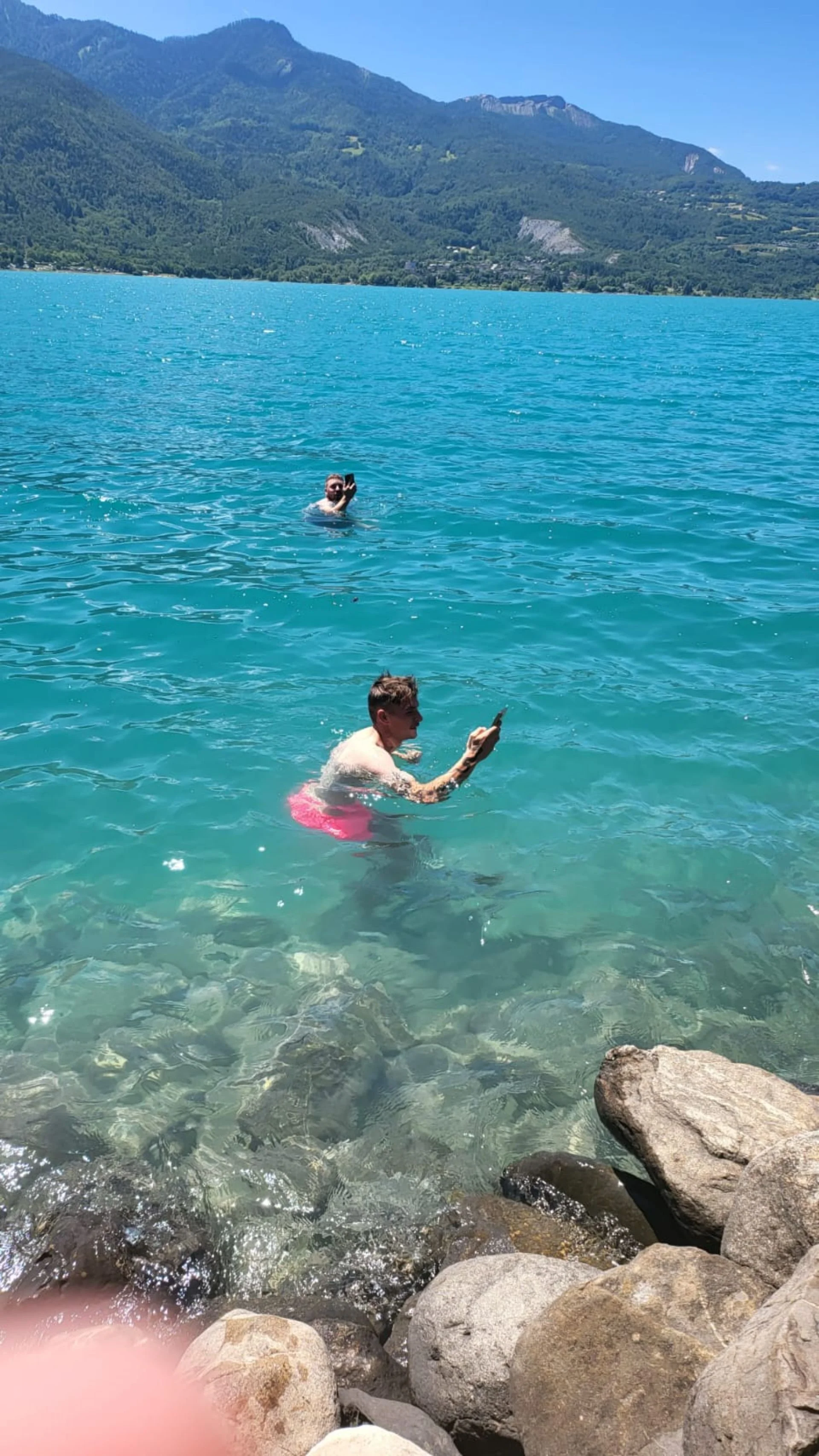 Two people swimming in a clear, turquoise lake with rocky shoreline, mountains and blue sky in the background.
