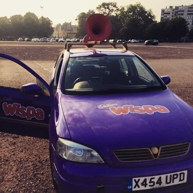 A purple car with Wispa logo is parked in an outdoor lot. The car has a large red horn mounted on its roof, and the driver's side door is open. The background shows other parked cars, trees, and buildings.