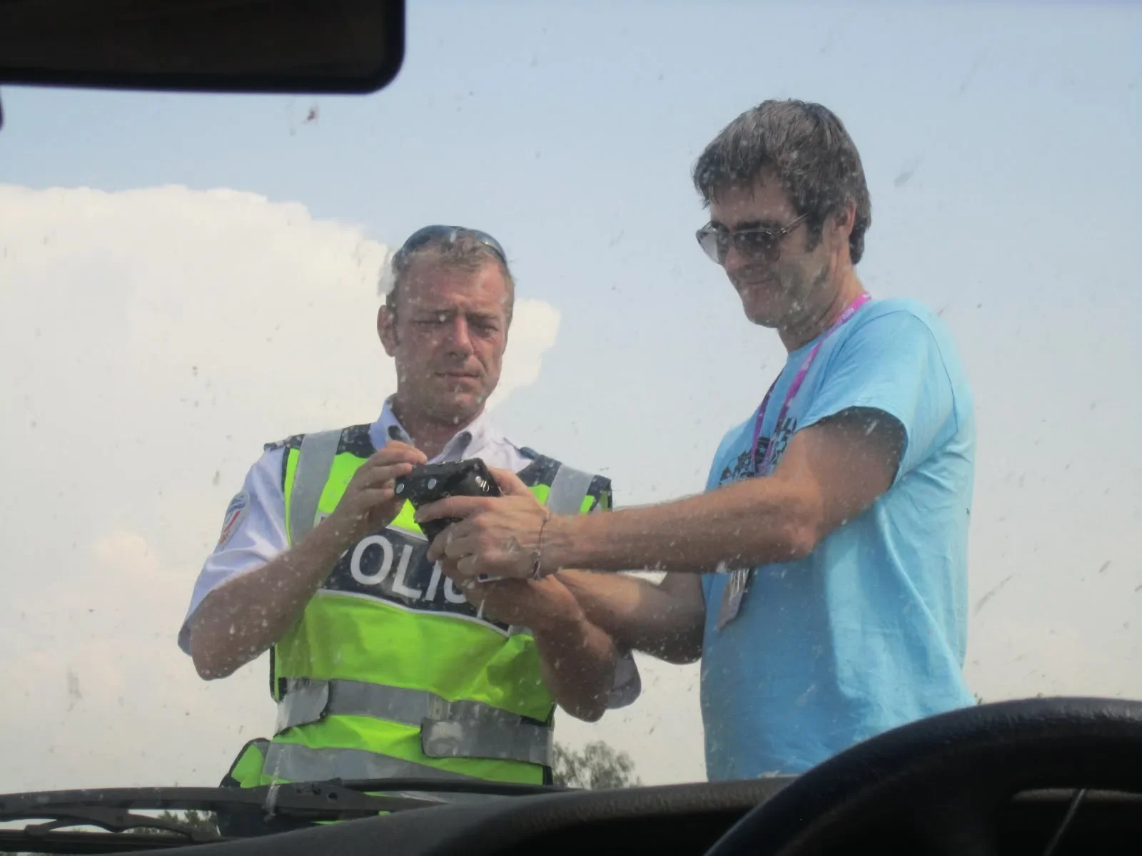 A police officer and a man in sunglasses are looking at a device together through the windshield of a vehicle, with a cloudy sky in the background.