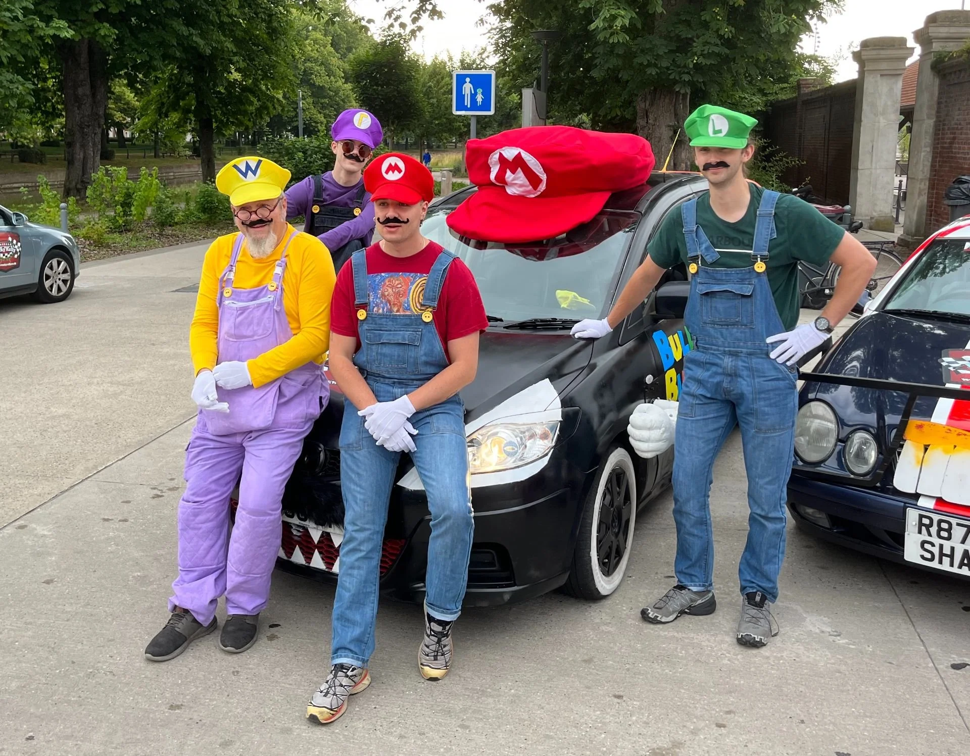 Five people dressed as Mario and Luigi characters from Super Mario Bros., with hats, mustaches, and gloves, standing beside a black car with a large red hat on the roof, in a parking lot with other cars and trees in the background.