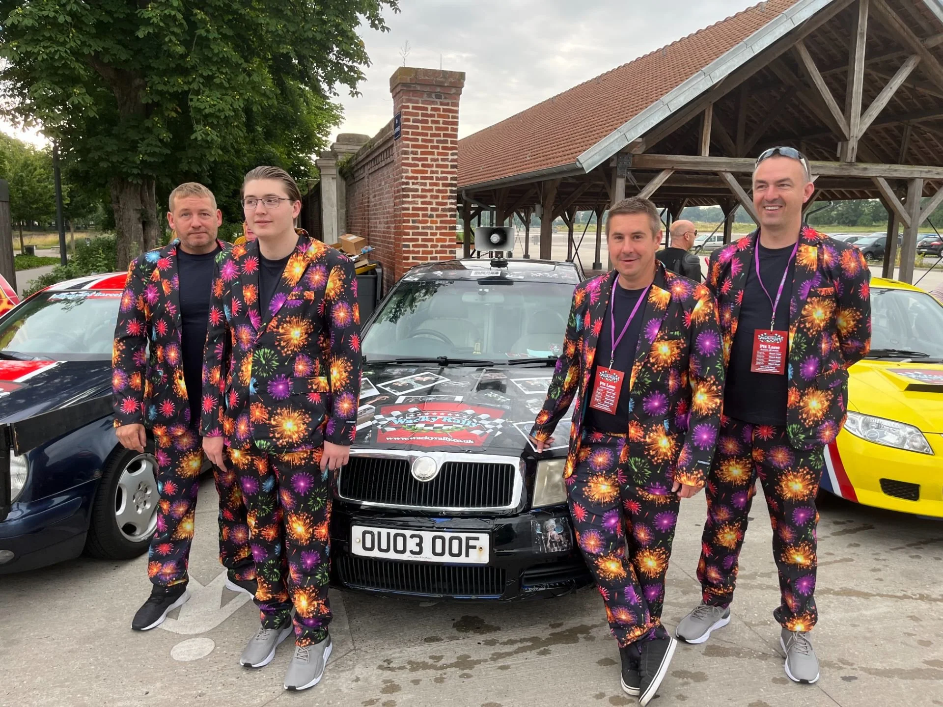 Four men dressed in matching black suits with colorful firework patterns stand in front of a race car at an outdoor event, with additional race cars and a wooden pavilion in the background.