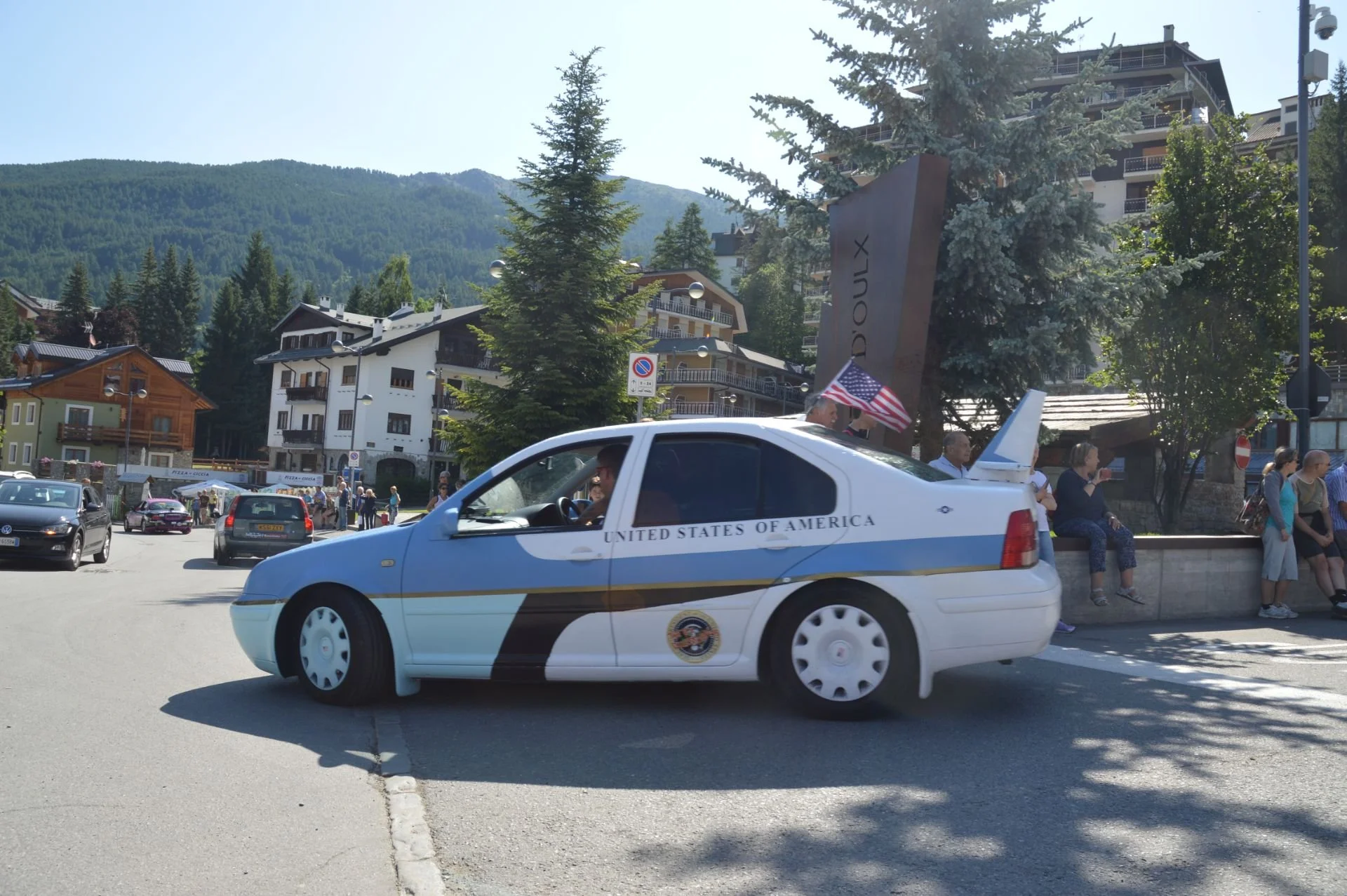 A car painted to resemble a U.S. government vehicle, labeled 'United States of America,' is driving through a mountain town with people sitting and standing on the sidewalk, and buildings and trees in the background.