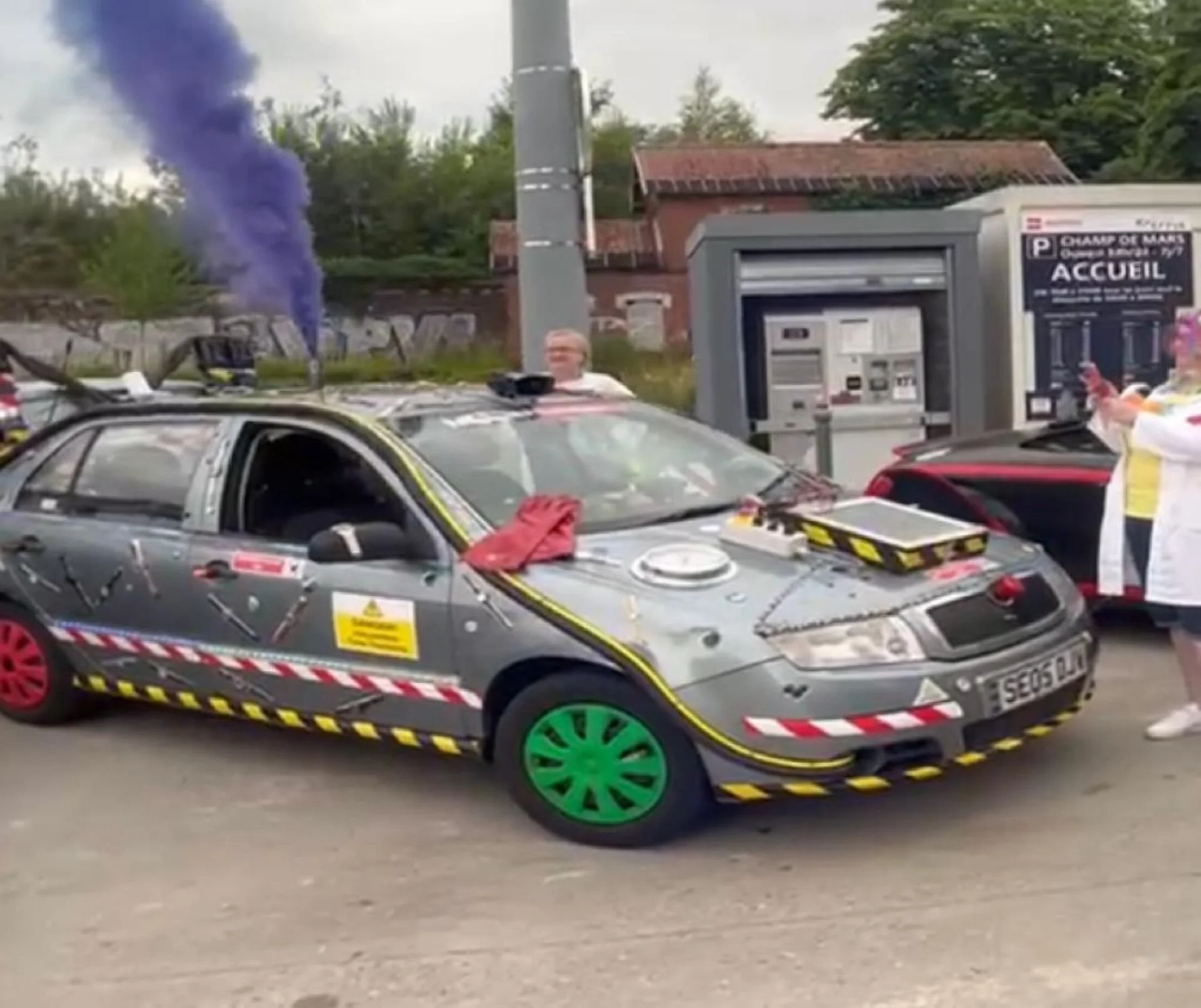 A crash test vehicle with warning stickers and colored wheel covers, emitting smoke, and two supervisors inspecting the vehicle at a testing site.