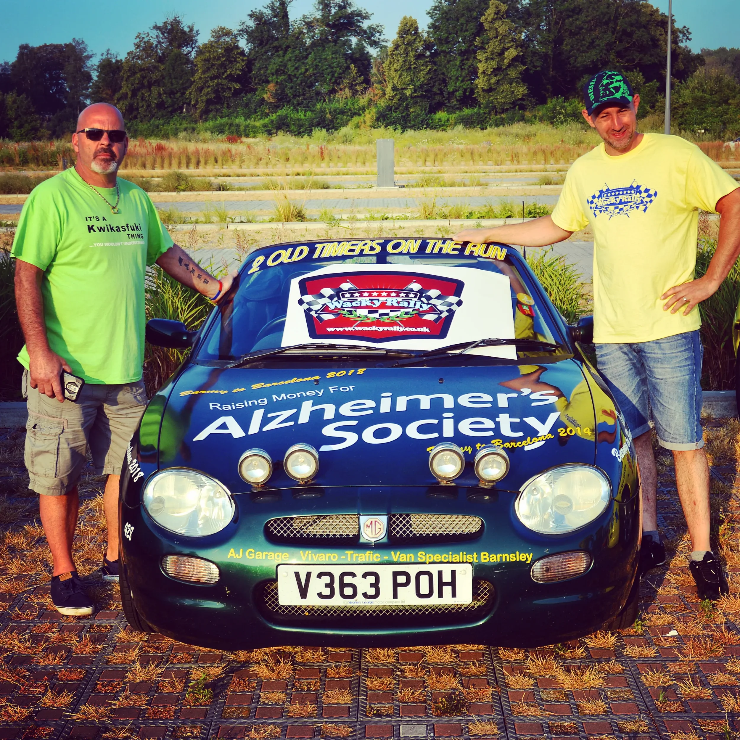 Two men standing beside a race car decorated with Alzheimer's Society logos and text, outdoors in a parking lot with trees in the background.