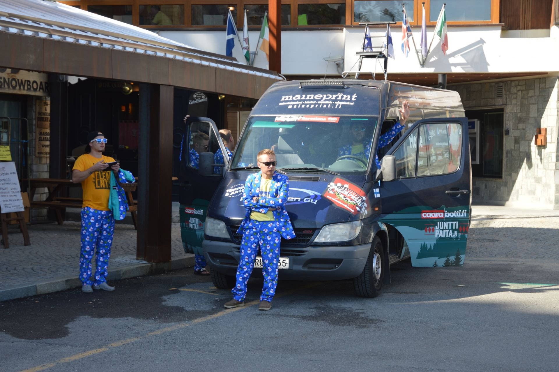Several people in matching blue pajama outfits standing and sitting near a black van with colorful logos, in front of a building with flags on the balcony.
