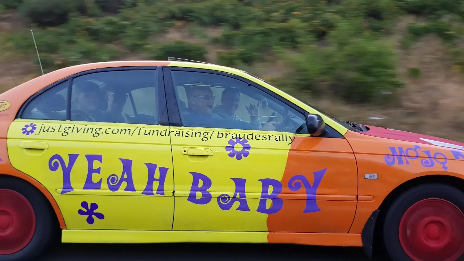 Colorful car with yellow and orange paint and purple text, promoting a fundraising website, with three people inside waving and driving on a road with greenery in the background.