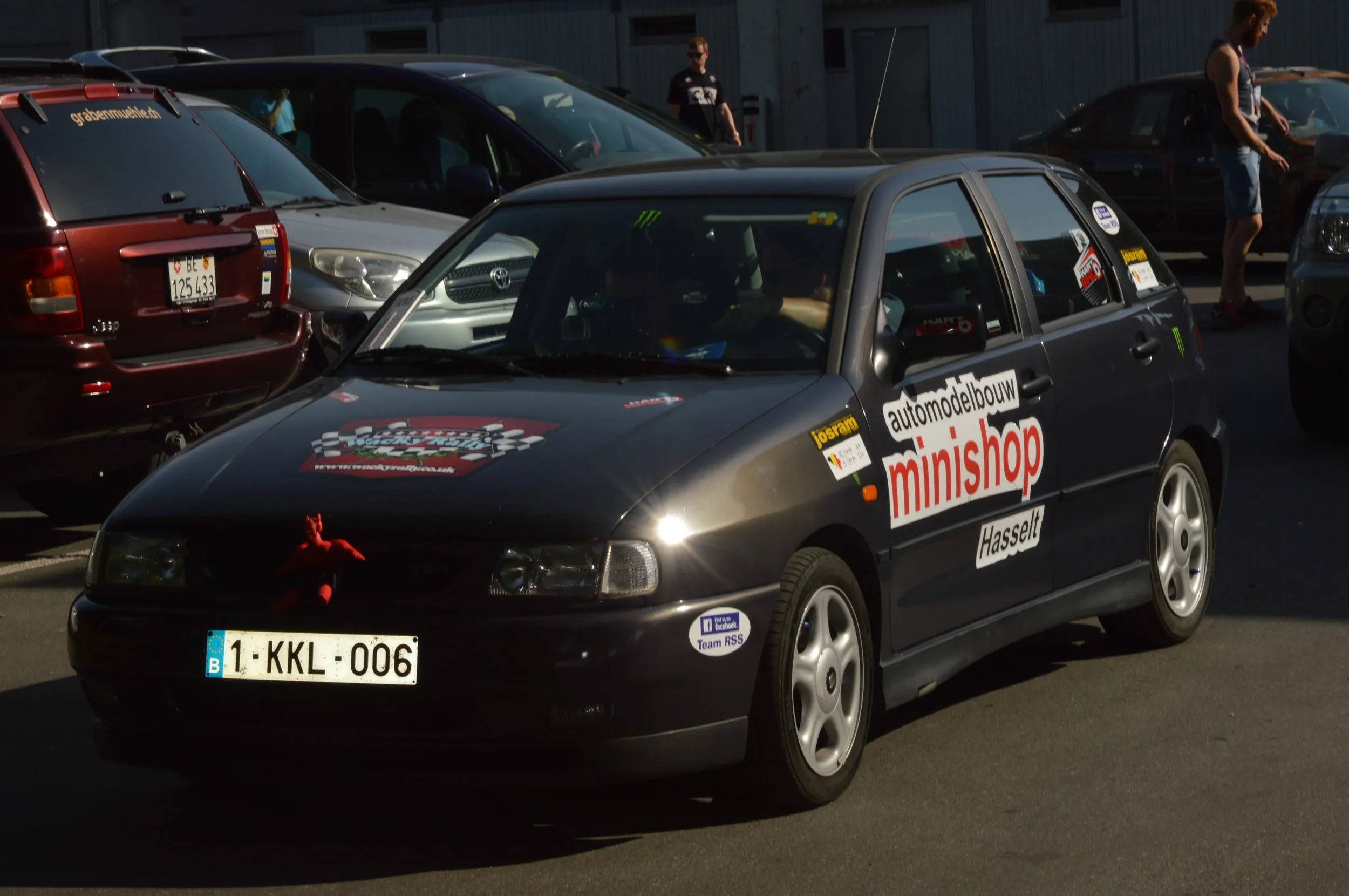 Black hatchback race car with sponsor decals parked among other vehicles, including a red SUV and a silver car, in a parking lot on a sunny day.