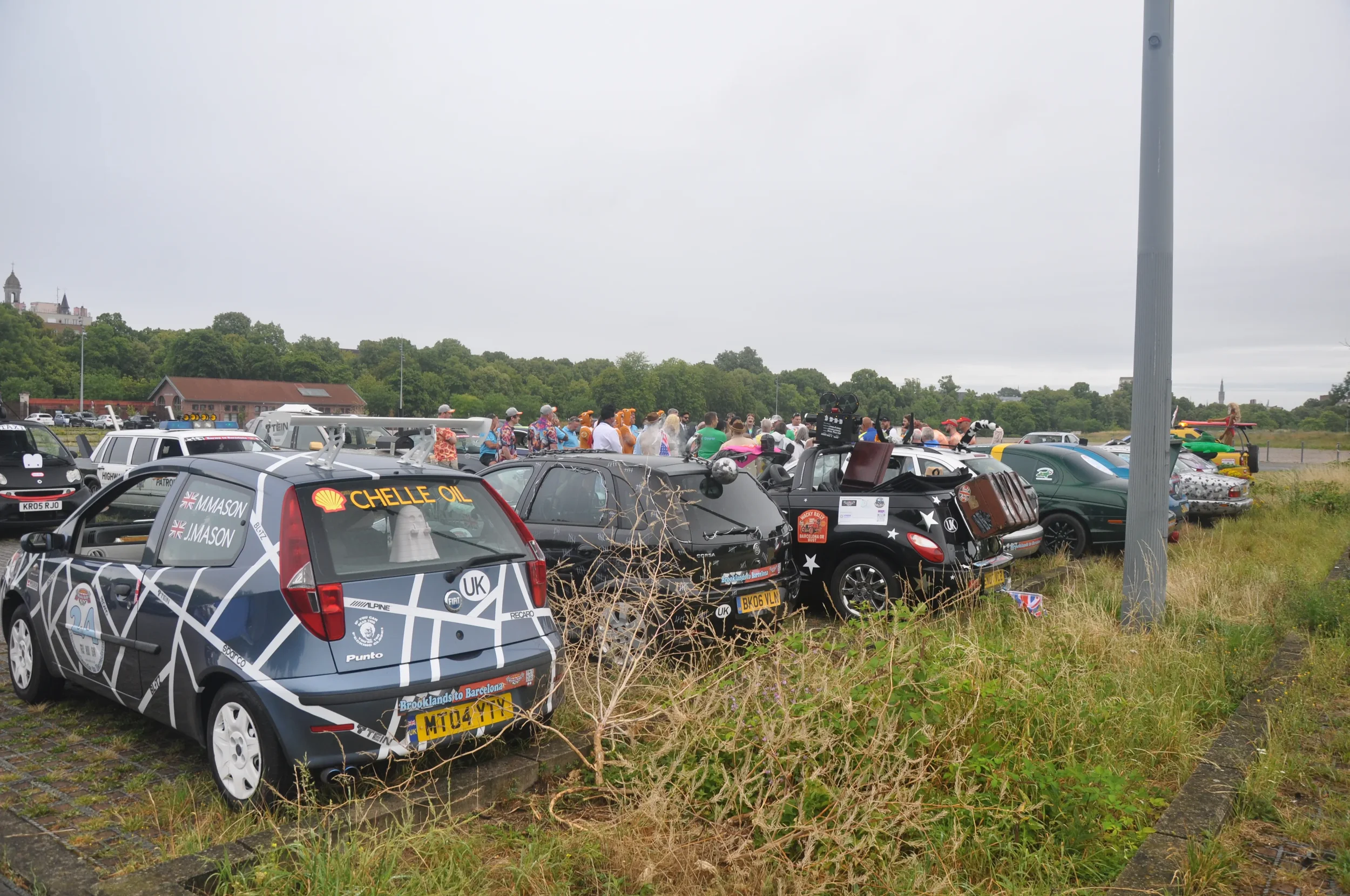 A row of parked cars, including a mini with racing decals and a small black race car, with a crowd of people and some aircraft in the background on an overcast day.