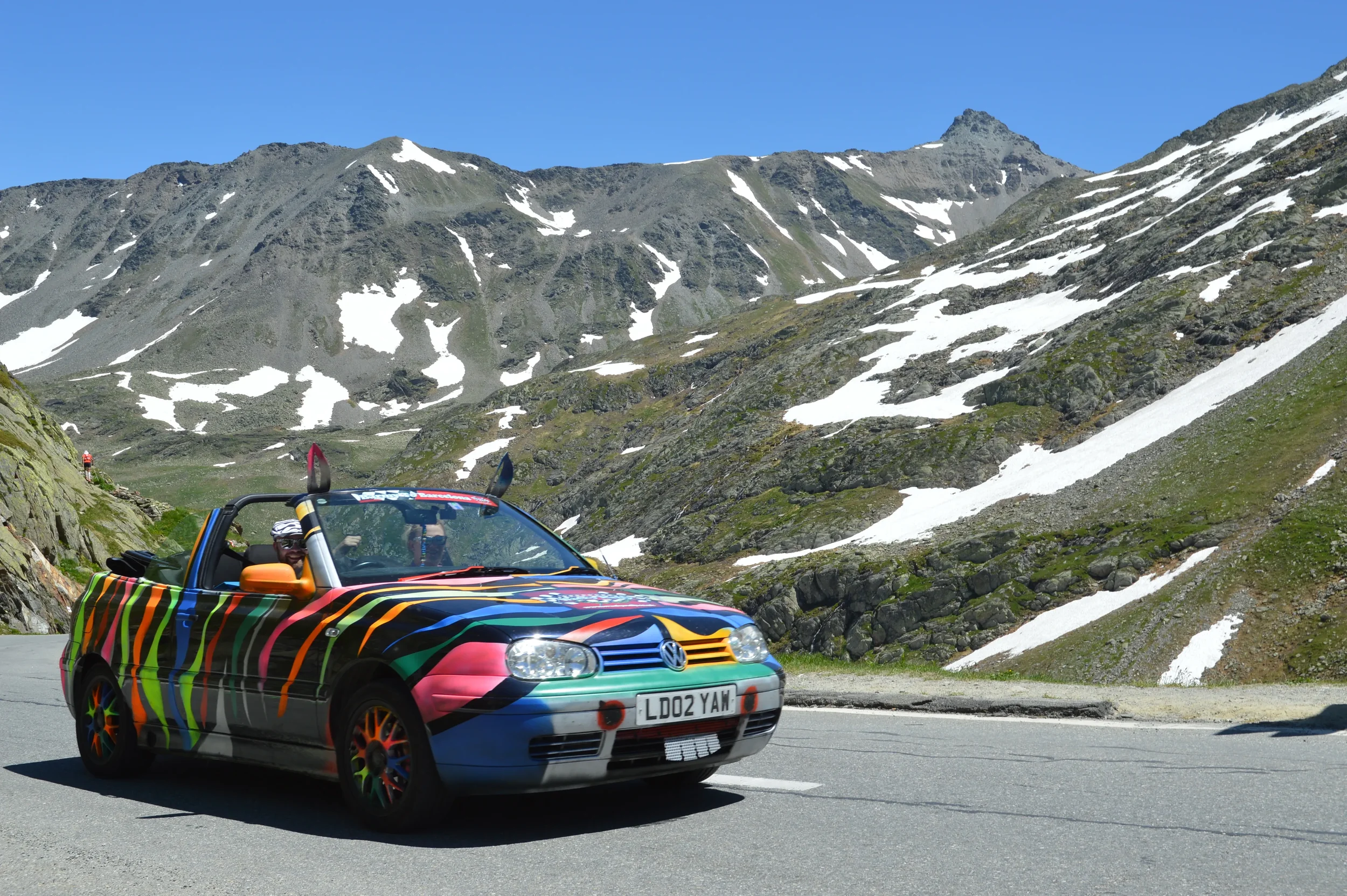 Colorful convertible car driving on mountain road with snow patches and rocky peaks in the background under a clear blue sky.