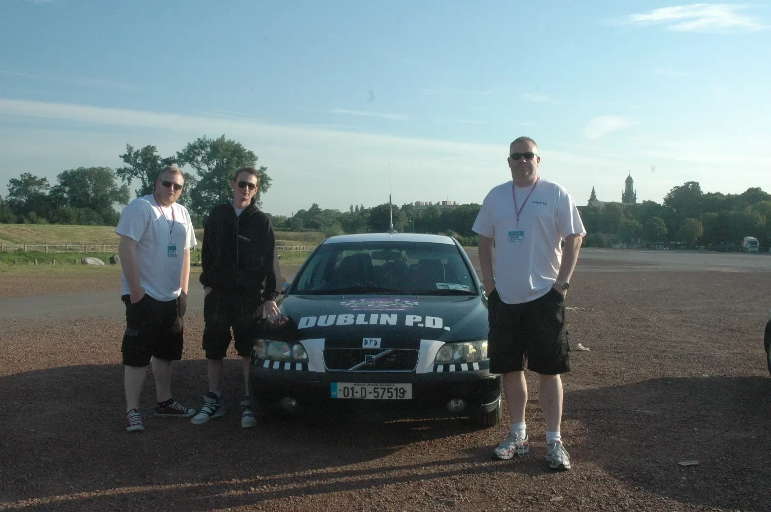 Three men standing near a black police car with Dublin P.D. written on it, in an outdoor setting with trees and a distant skyline in the background, during daylight.