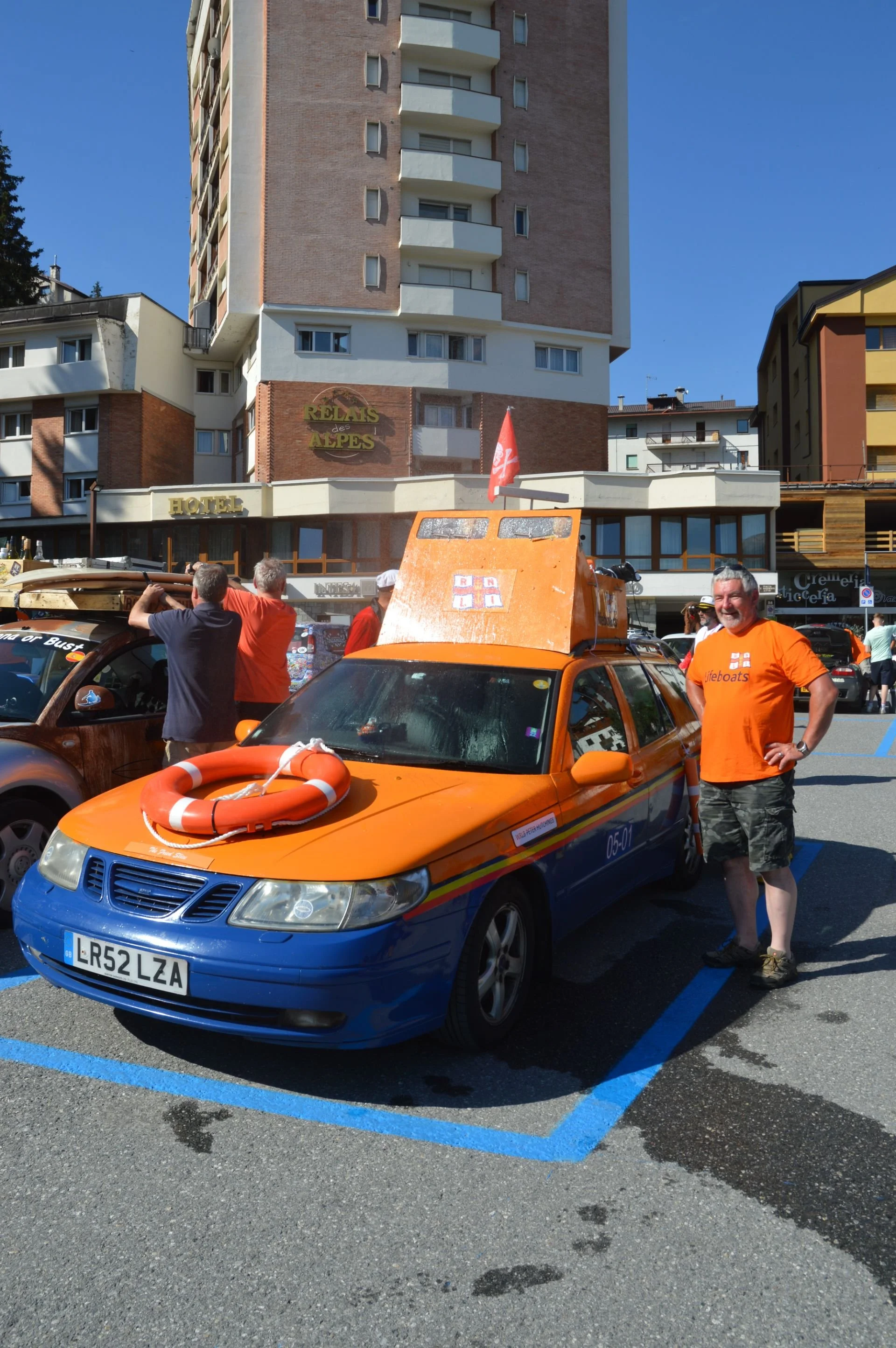 A man standing next to a colorful car decorated for a parade or event, holding a flotation device on the hood of the car. The car has a lifebuoy on its hood and a small boat on top. In the background are other people, buildings, and a hotel sign, wit