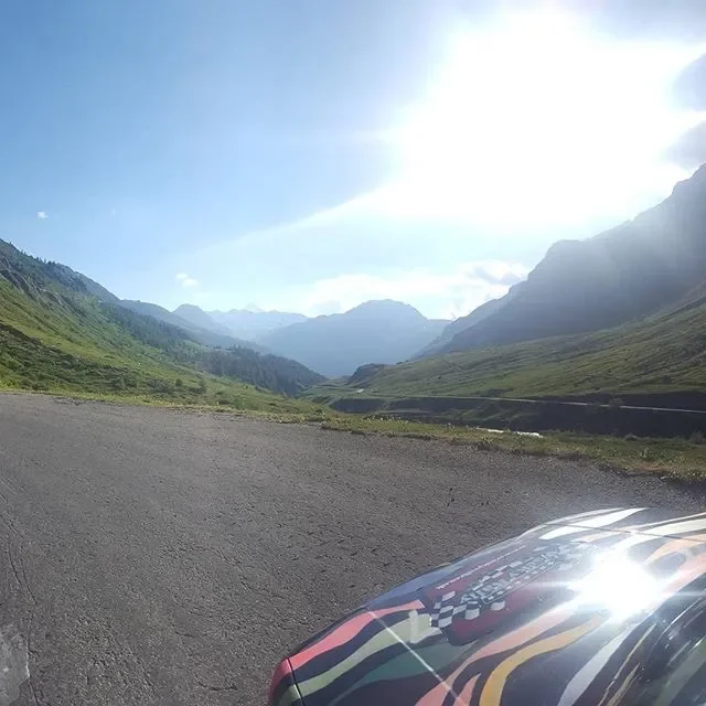 A scenic mountain landscape with green hills, distant mountains, a cloudy sky, and a portion of a race car with a checkered flag pattern in the foreground.