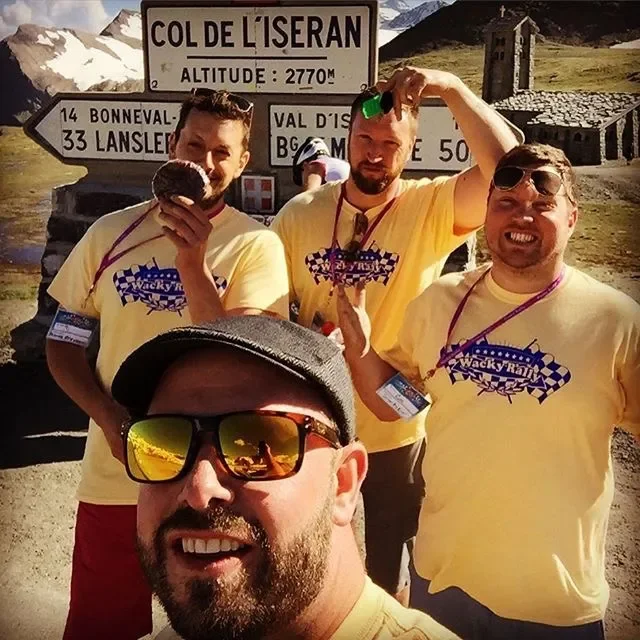 Four men in yellow t-shirts with 'Wacky Ral' logos, one taking a selfie, standing in front of a mountain road sign at Col de l'Iseran, with snowy peaks in the background.