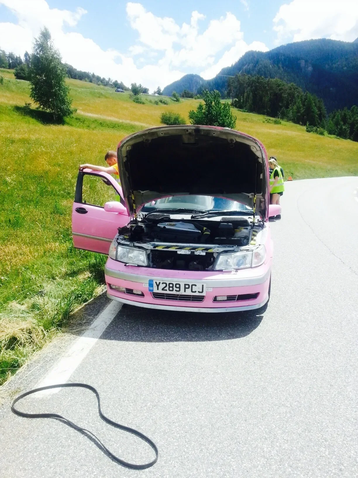 A pink car with open hood and driver-side door stopped on the side of a countryside road. Two people, a boy and a woman, are inspecting the car; the boy is leaning on the door, and the woman is standing behind the car wearing a safety vest. The scene